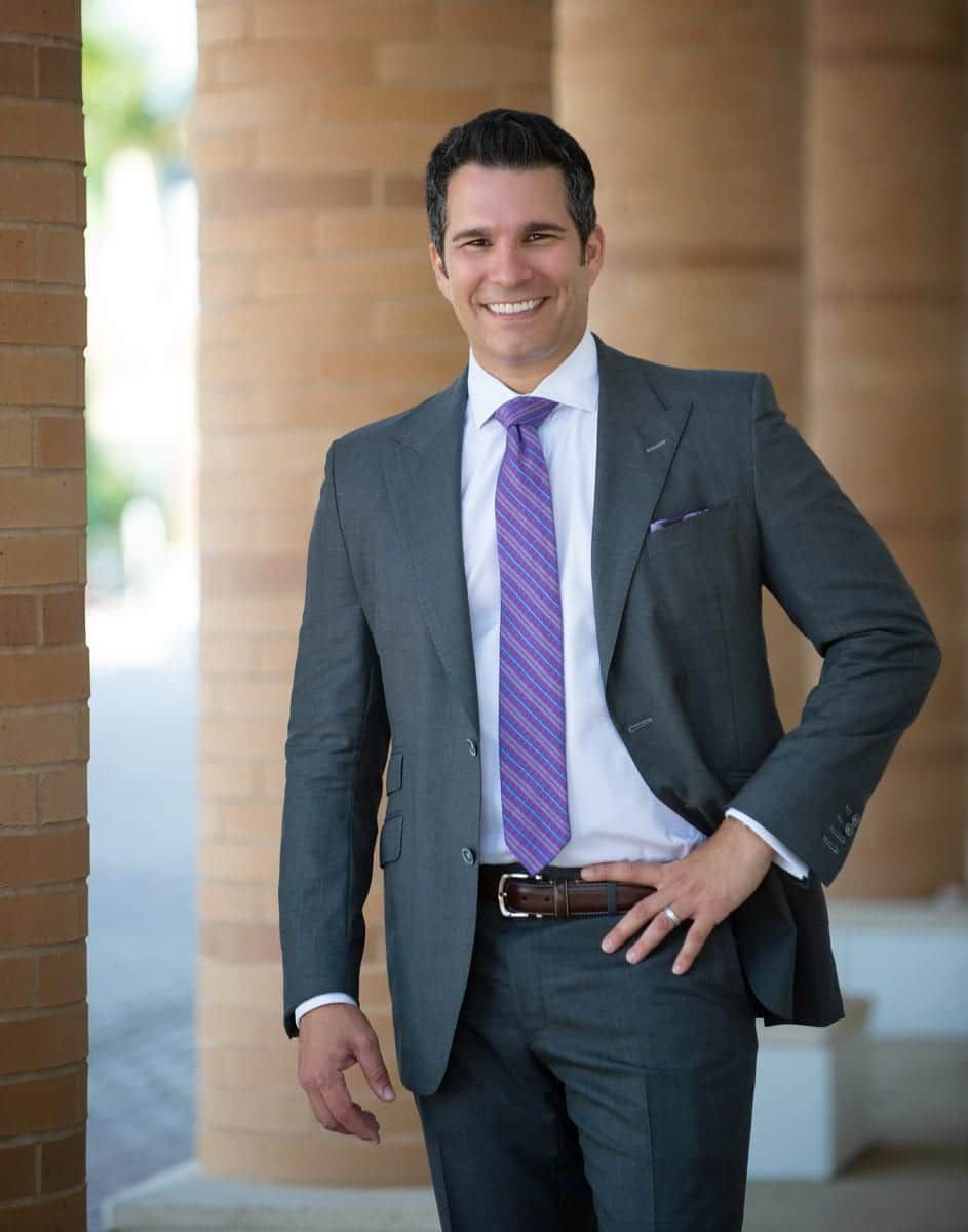 Man in gray suit with purple tie smiling, hand on hip, outdoors near pillars.