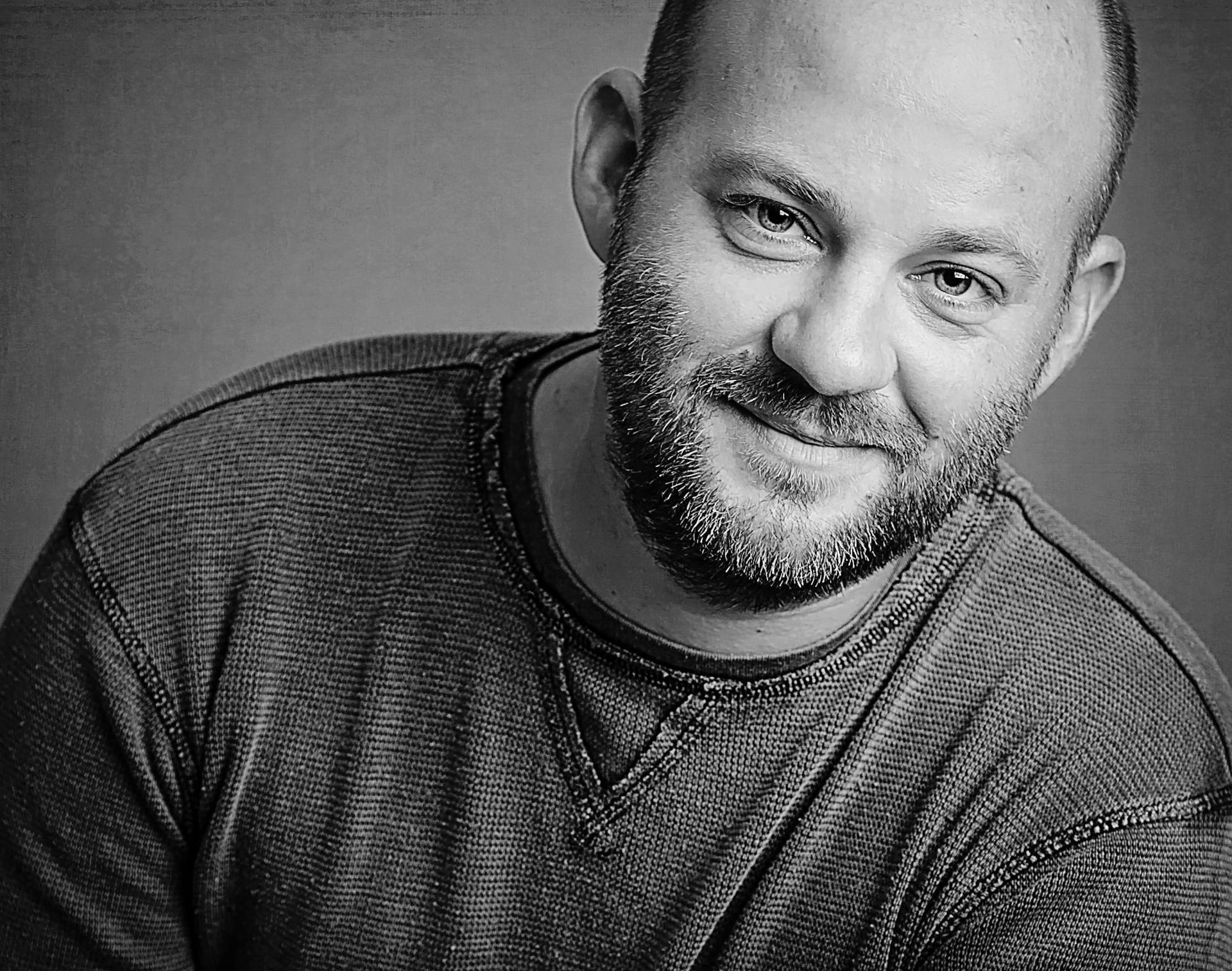 Man with a shaved head and beard smiles, wearing a dark crew neck sweater.