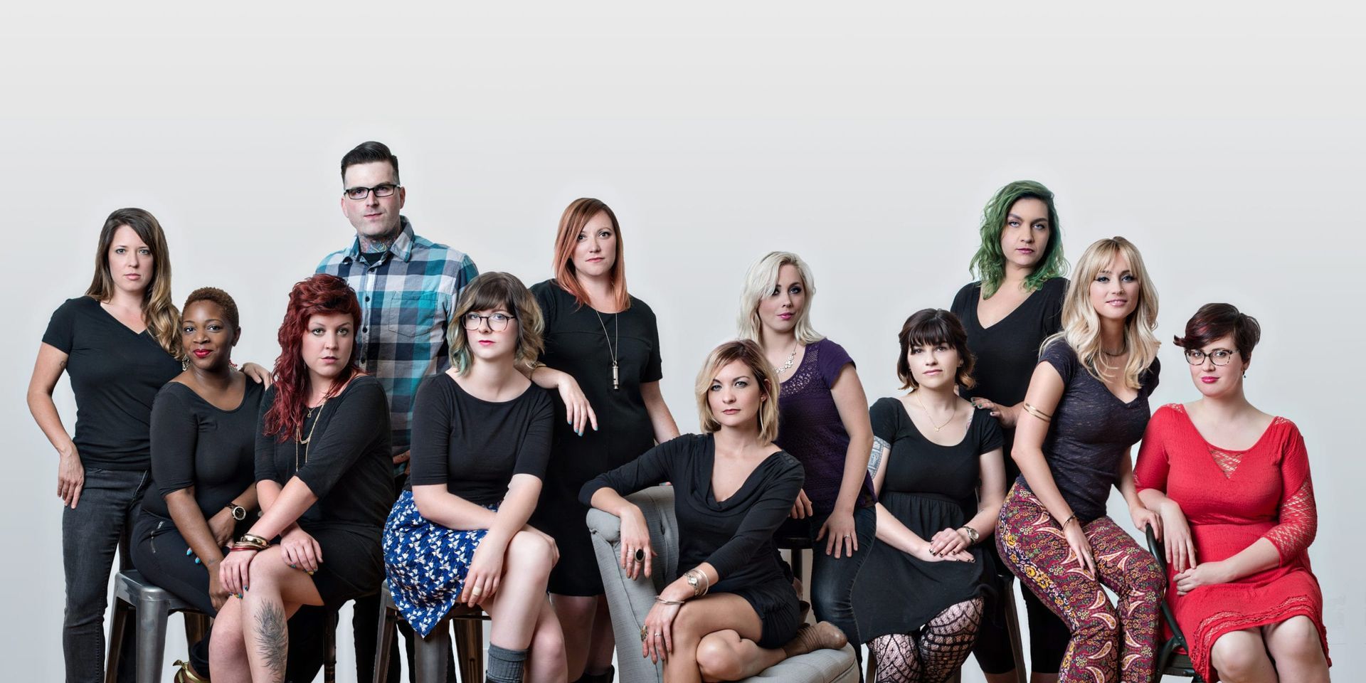 Group of diverse people posing for a photo, many in black dresses, some seated, against a white background.