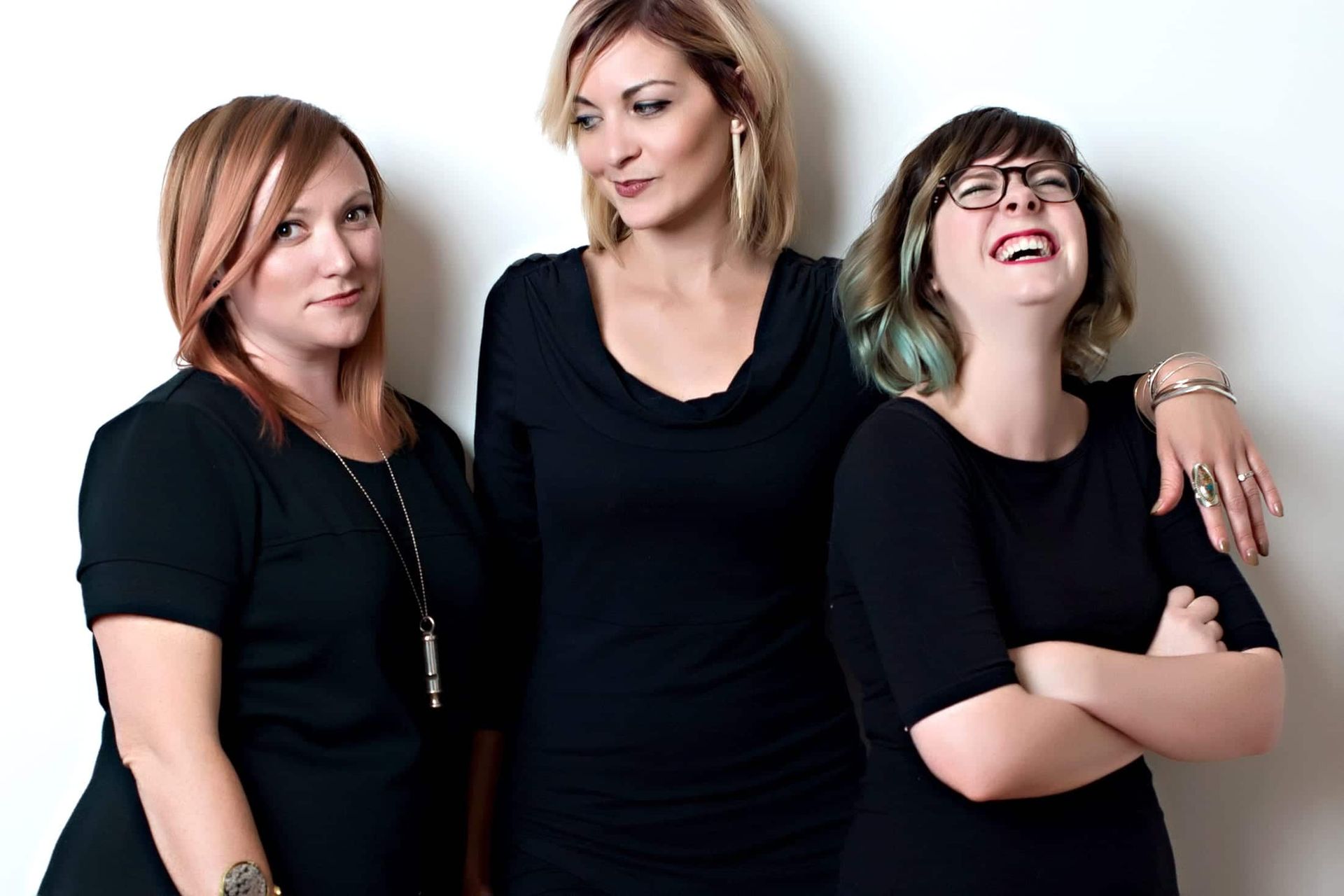 Three women in black clothing pose; one laughs, one smirks, one with a playful expression.