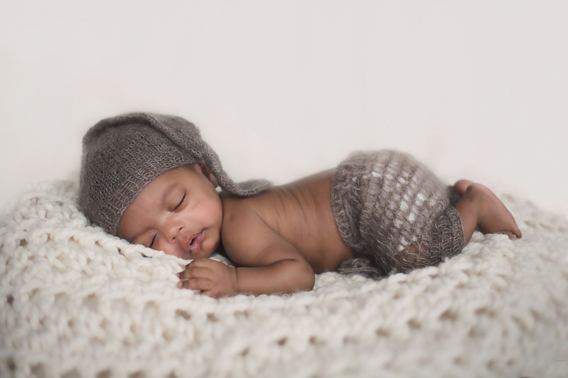 Newborn baby asleep on a cream knitted blanket, wearing a gray cap and shorts.