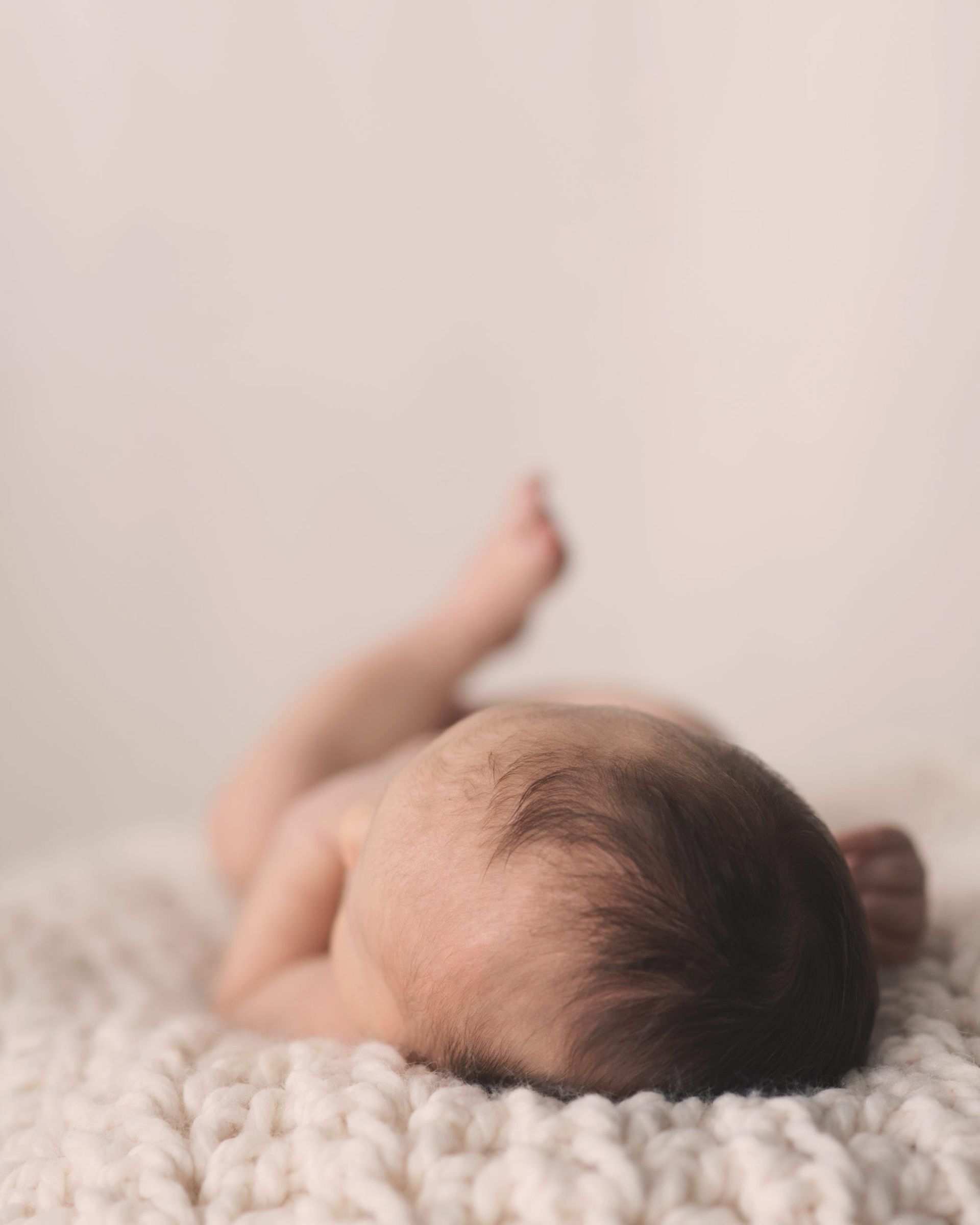 Newborn baby lying on a cream-colored knitted blanket; back and head are visible.