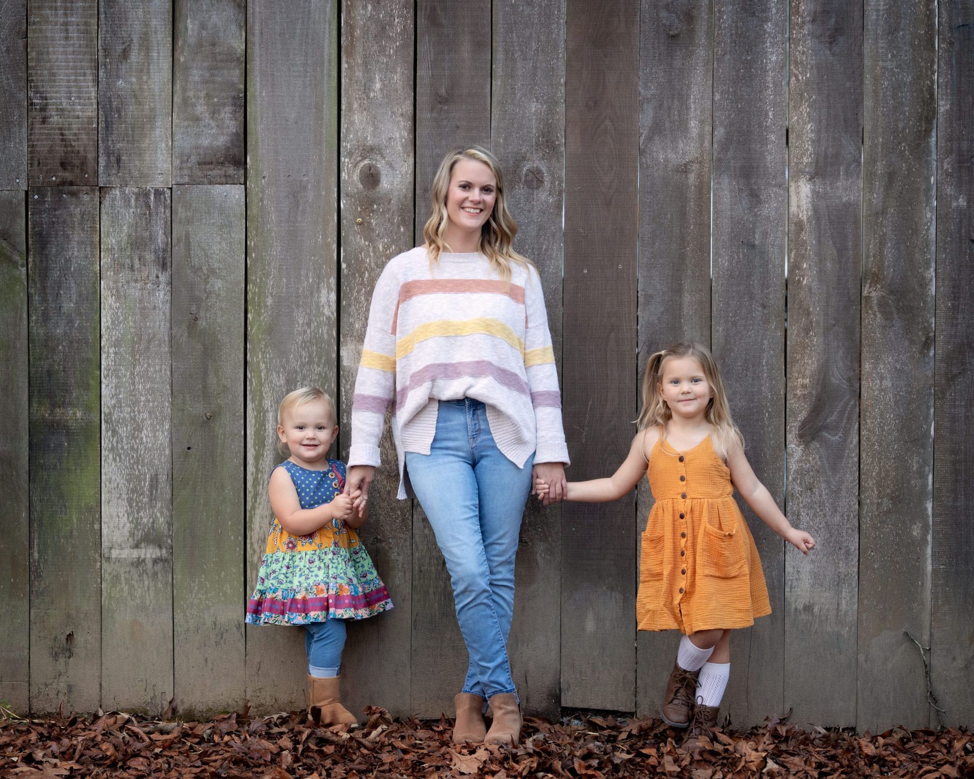 Woman and two young girls smiling, holding hands, in front of a weathered wooden wall.