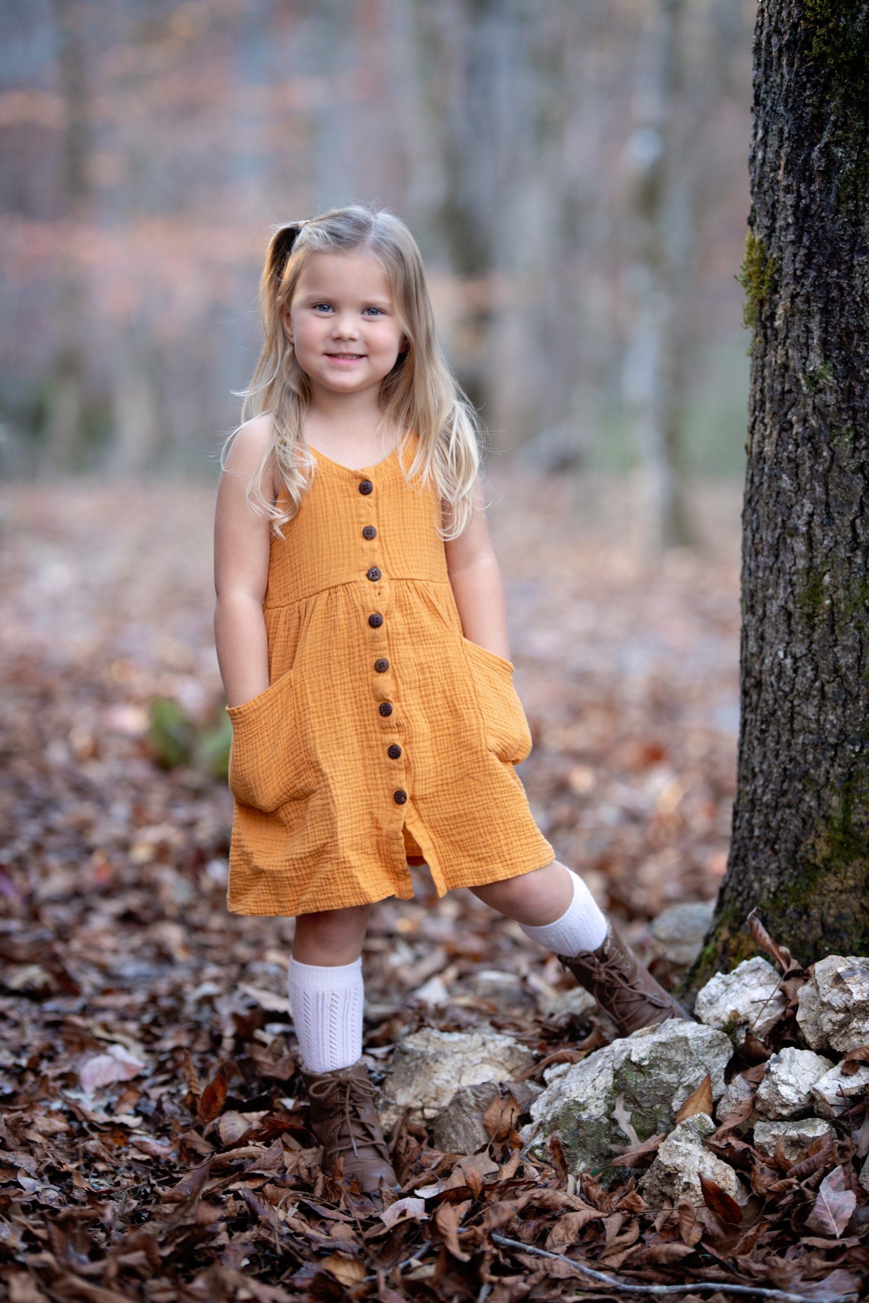 Young girl in mustard dress, smiling, standing near tree in outdoor setting.