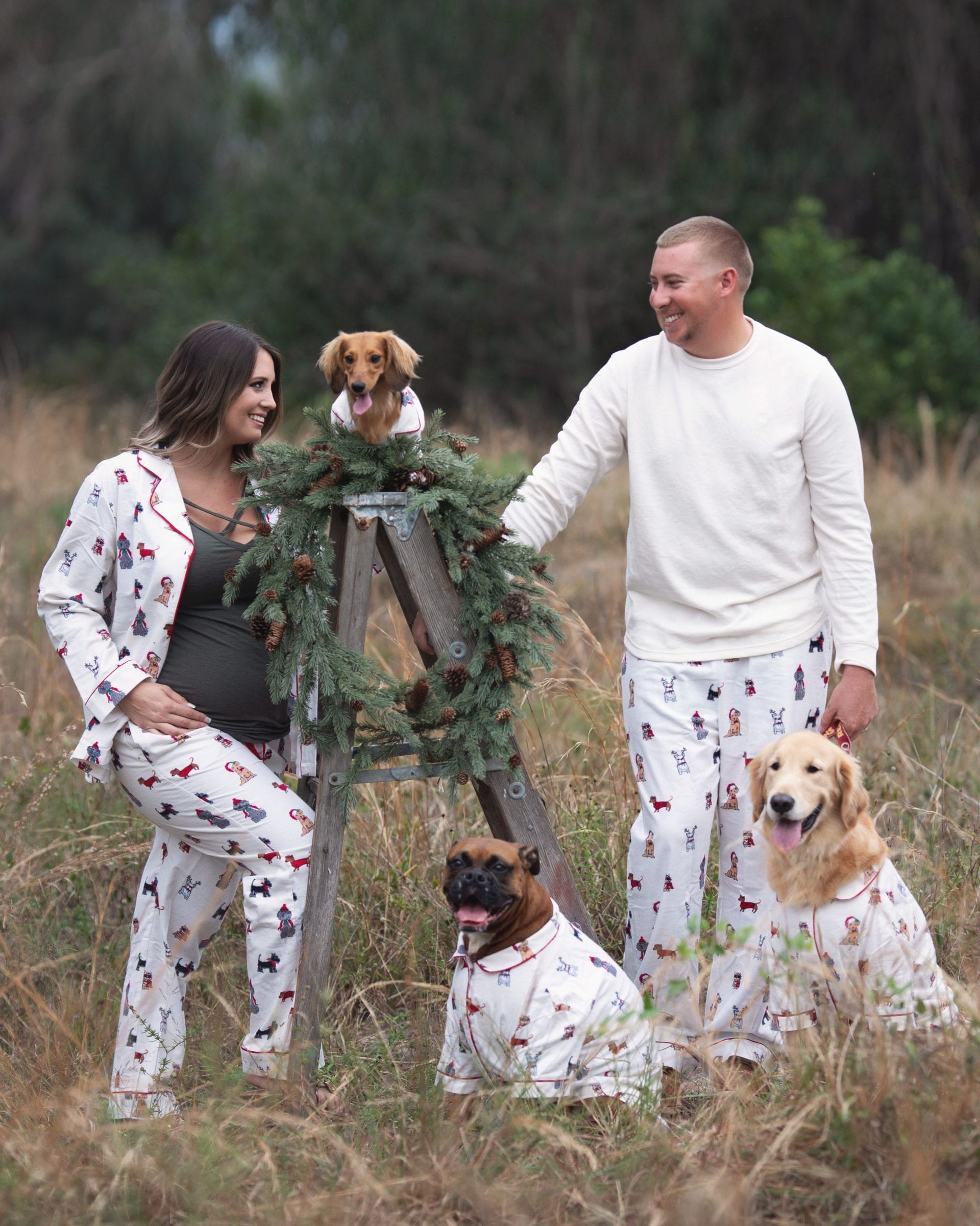 Pregnant couple and dogs in matching pajamas outdoors, smiling, near a decorated ladder.