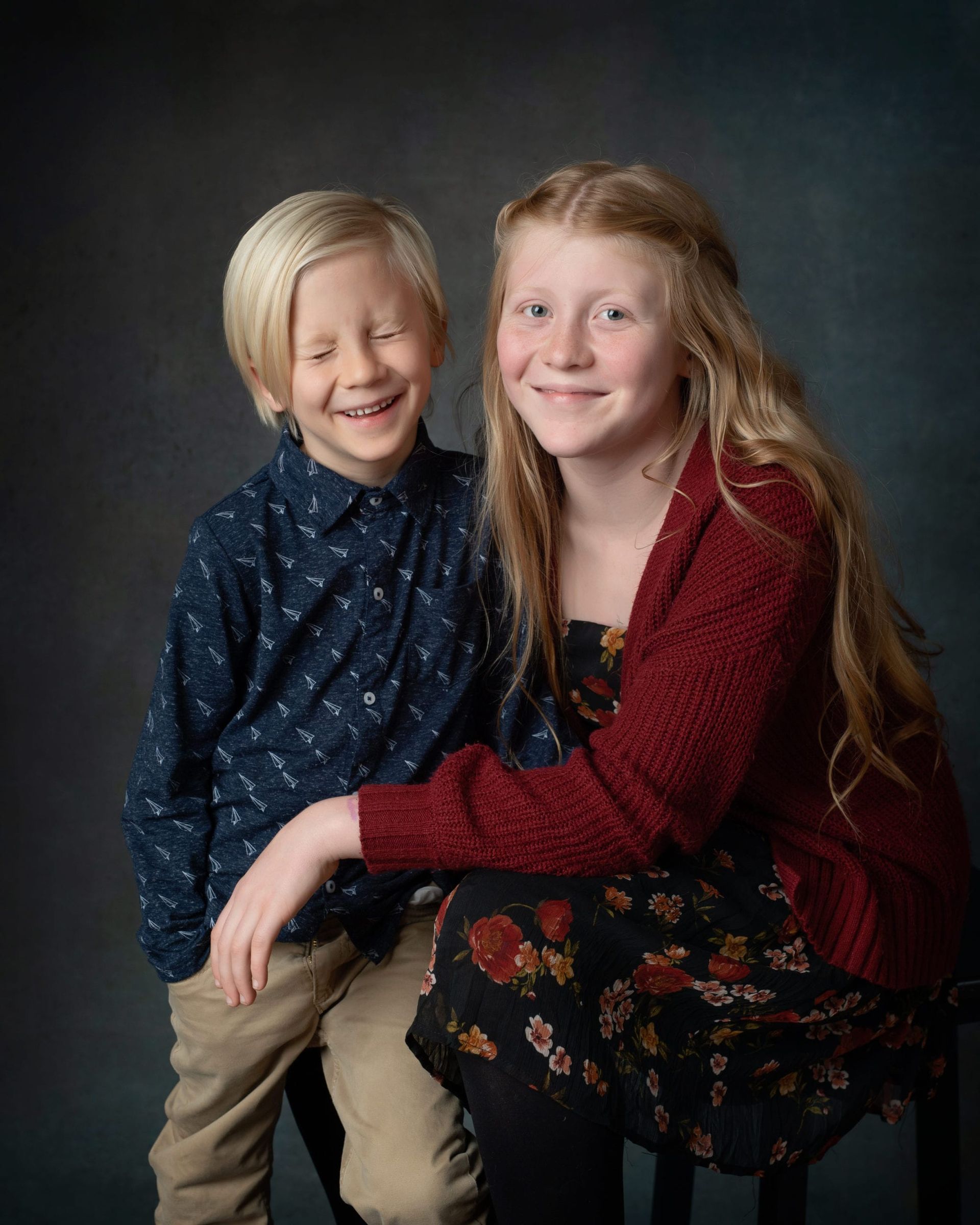 Boy with blond hair laughing with sister, both smiling, against a dark backdrop.