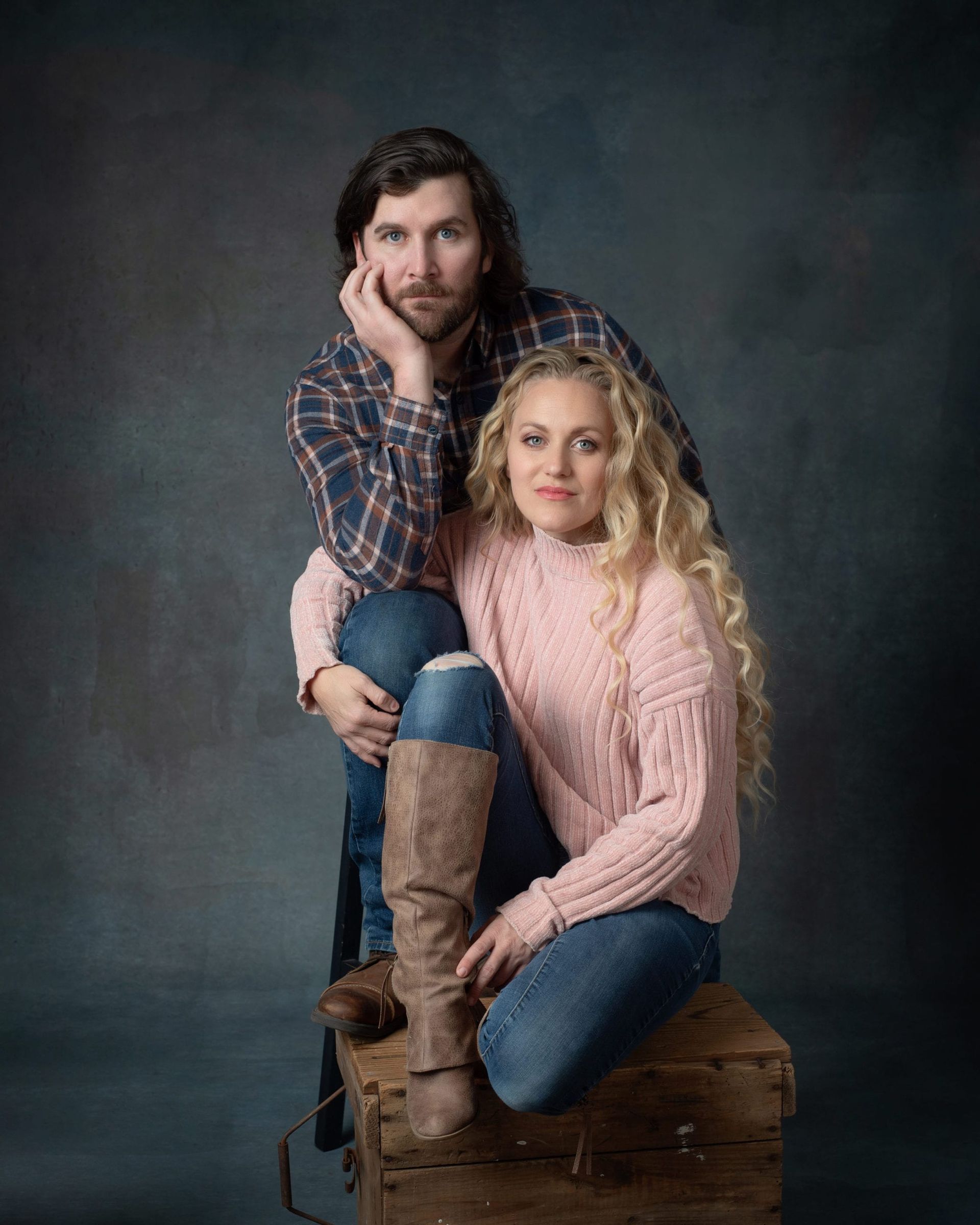 Couple posing on a box; woman in pink sweater, man in flannel shirt, against a dark backdrop.