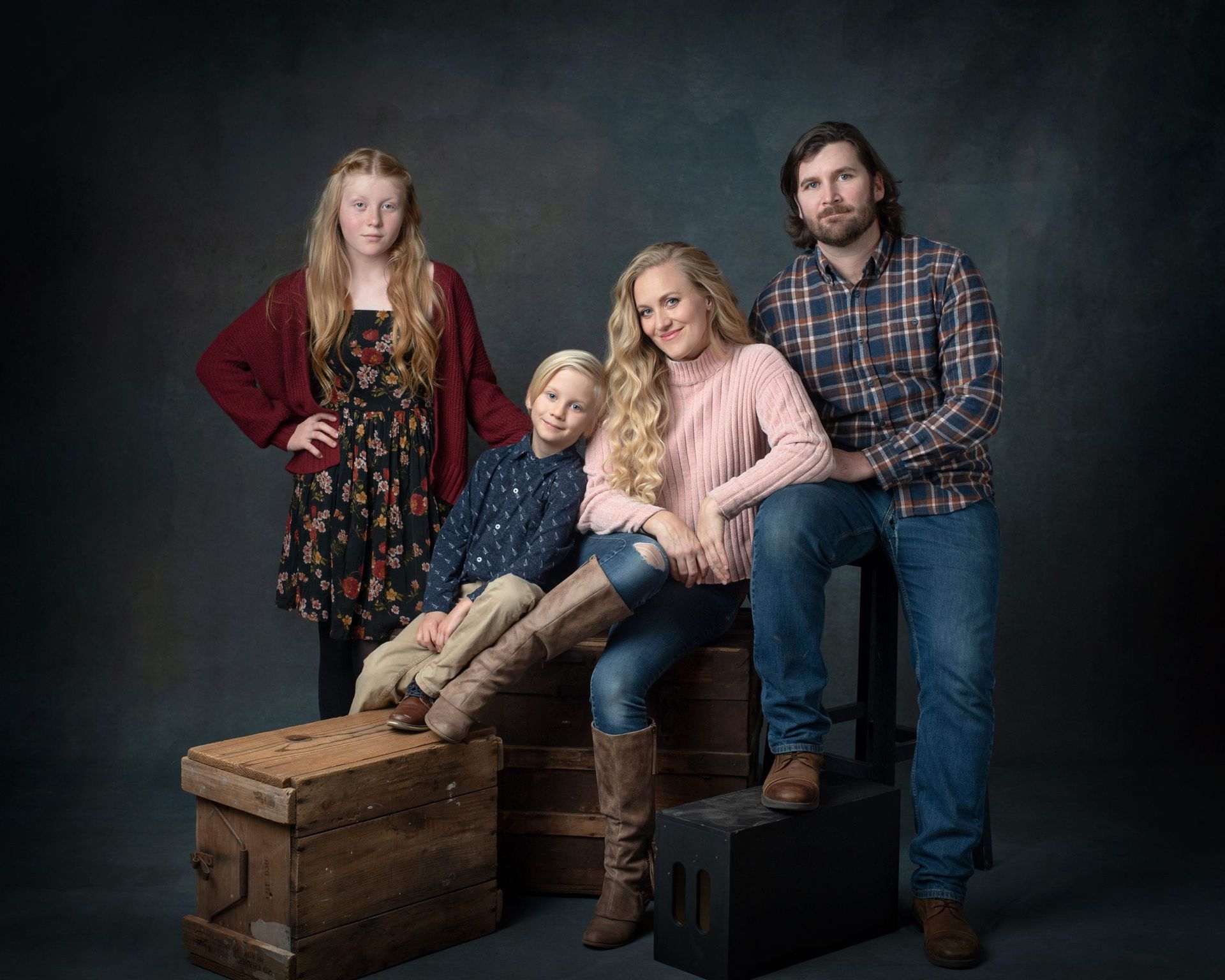 Family portrait: Two adults, two children, posed on boxes against a dark background. Blonde woman, man, boy. Teenage girl.