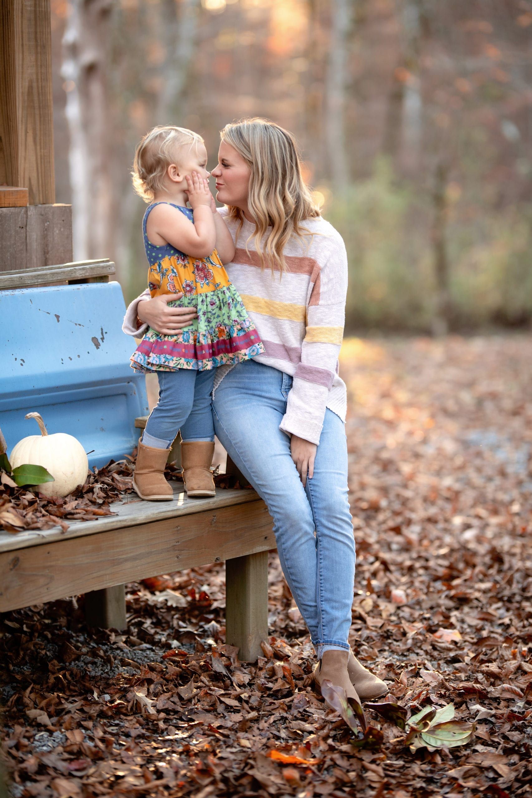 Mother whispers to child outdoors; fall leaves and pumpkin, sunlit background.