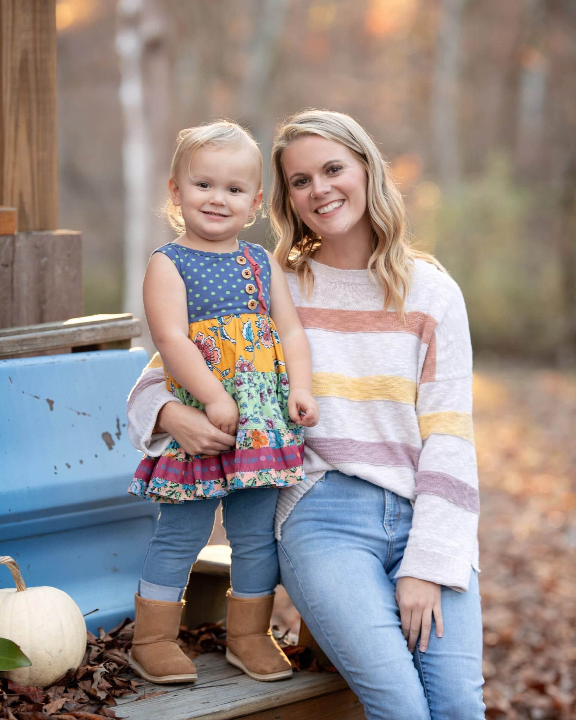 Woman smiles, holding a toddler. They are outdoors in autumn, near a pumpkin.