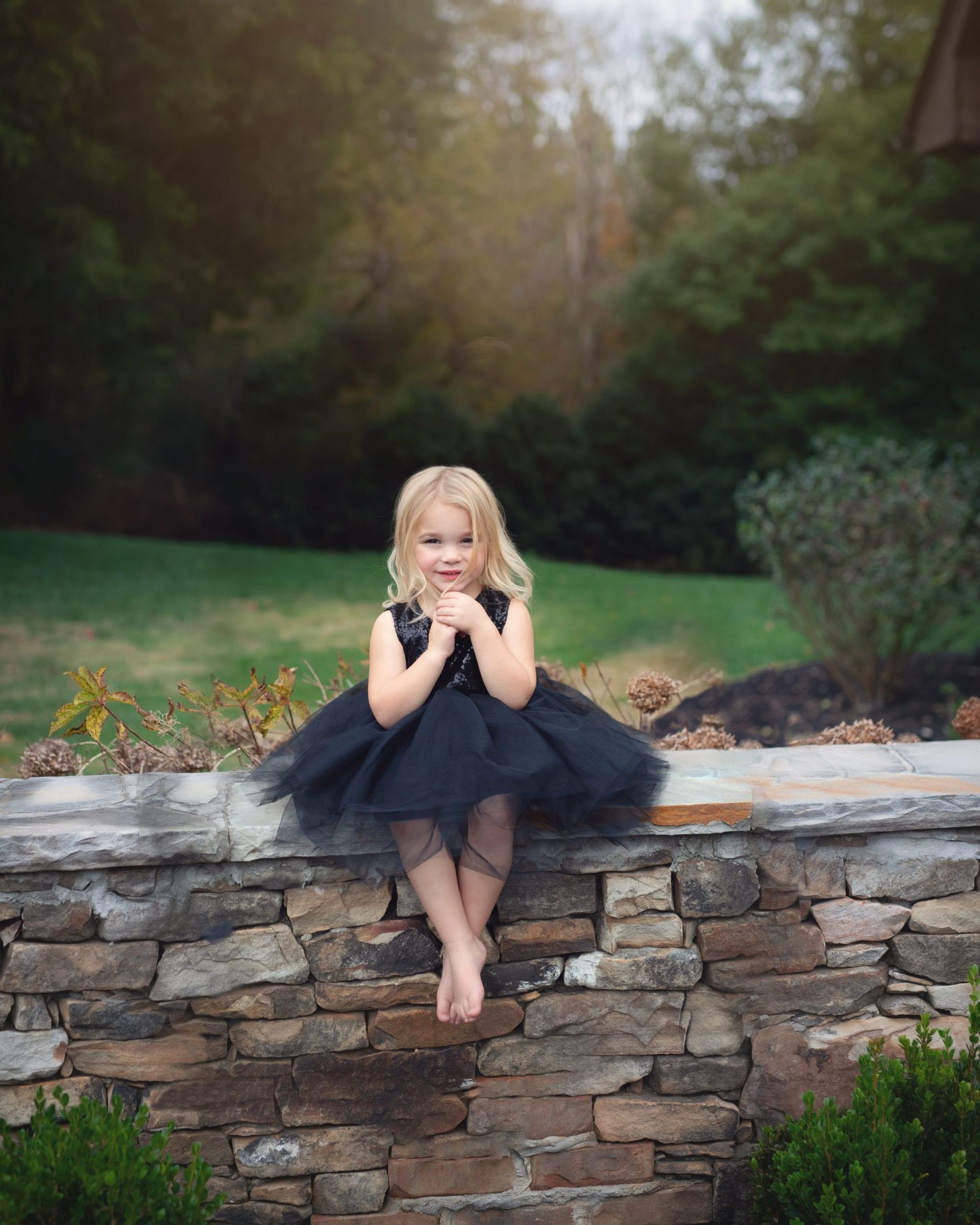 Blonde girl in black dress sits on stone wall, looking at camera. Green grass and trees in background.