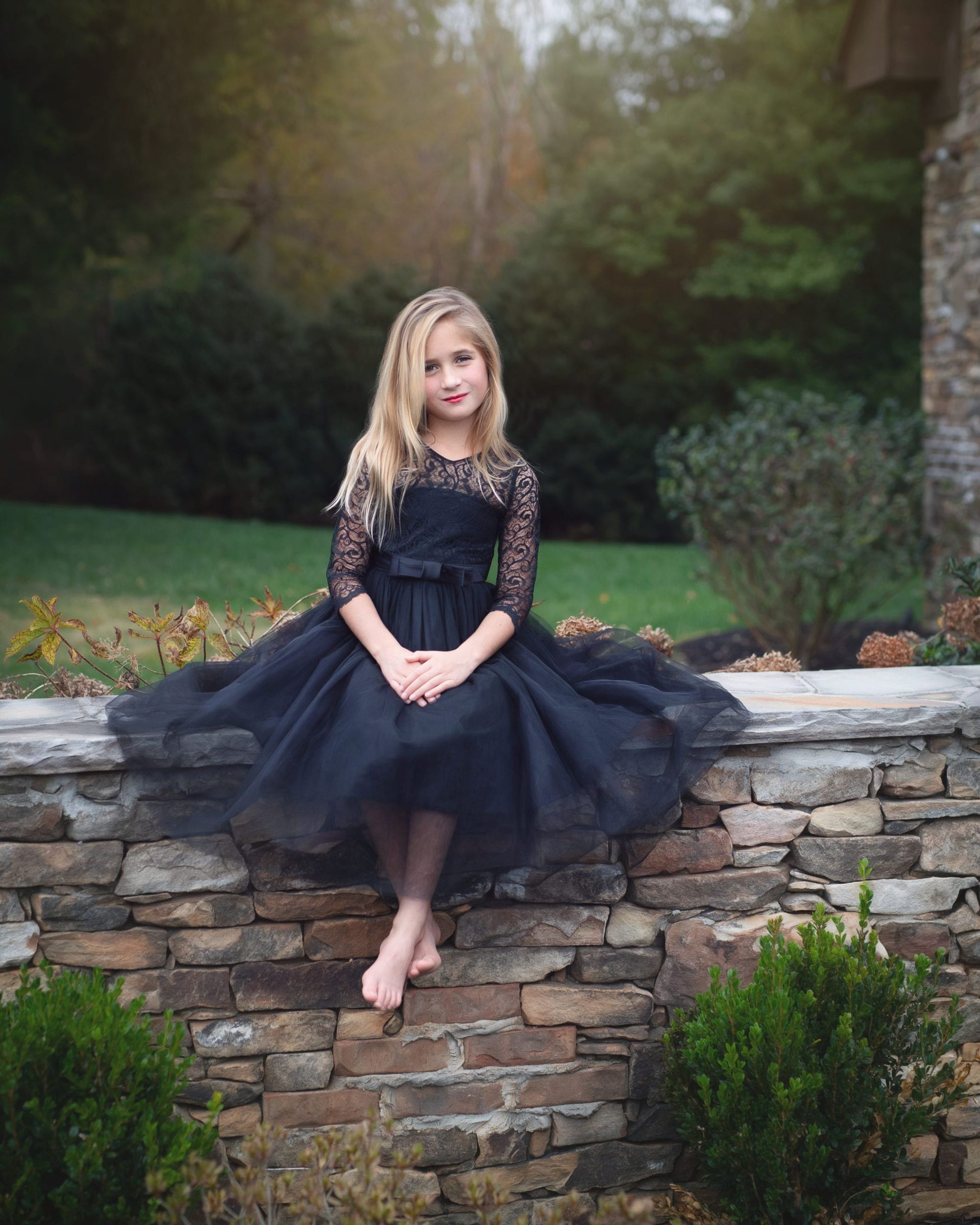 Young girl with blonde hair in black dress sits barefoot on a stone wall outdoors.