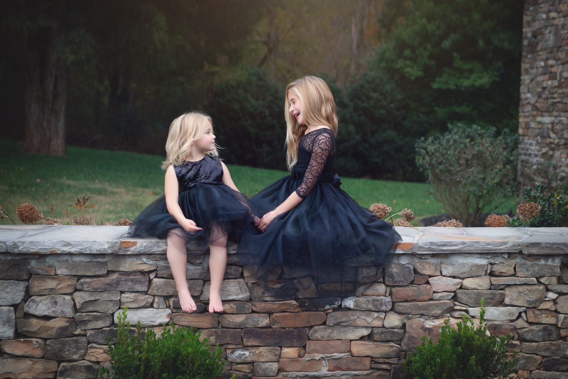 Two young girls in black dresses holding hands, sitting on a stone wall in a garden.