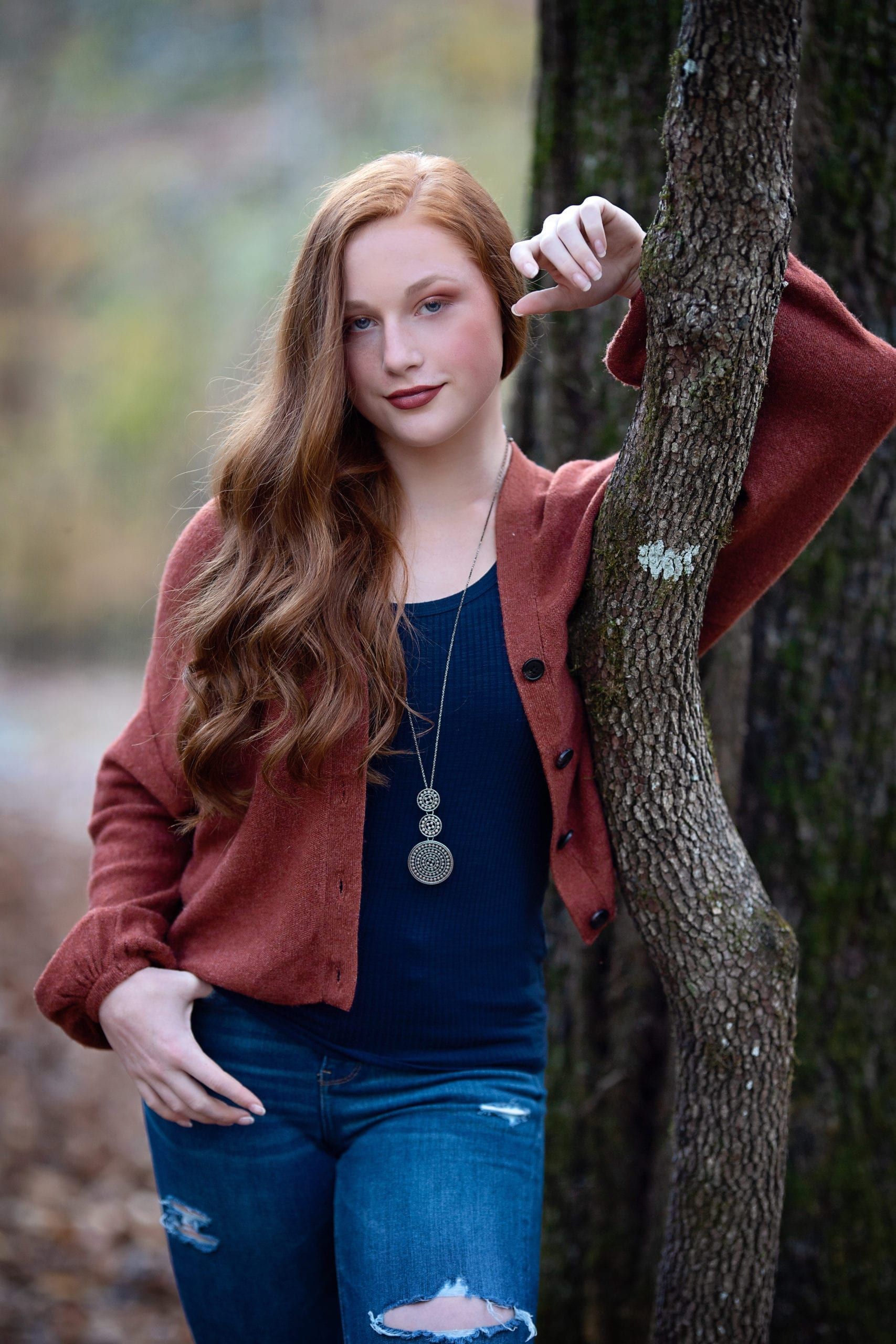 Red-haired woman in jeans and cardigan leans on a tree in a wooded setting.