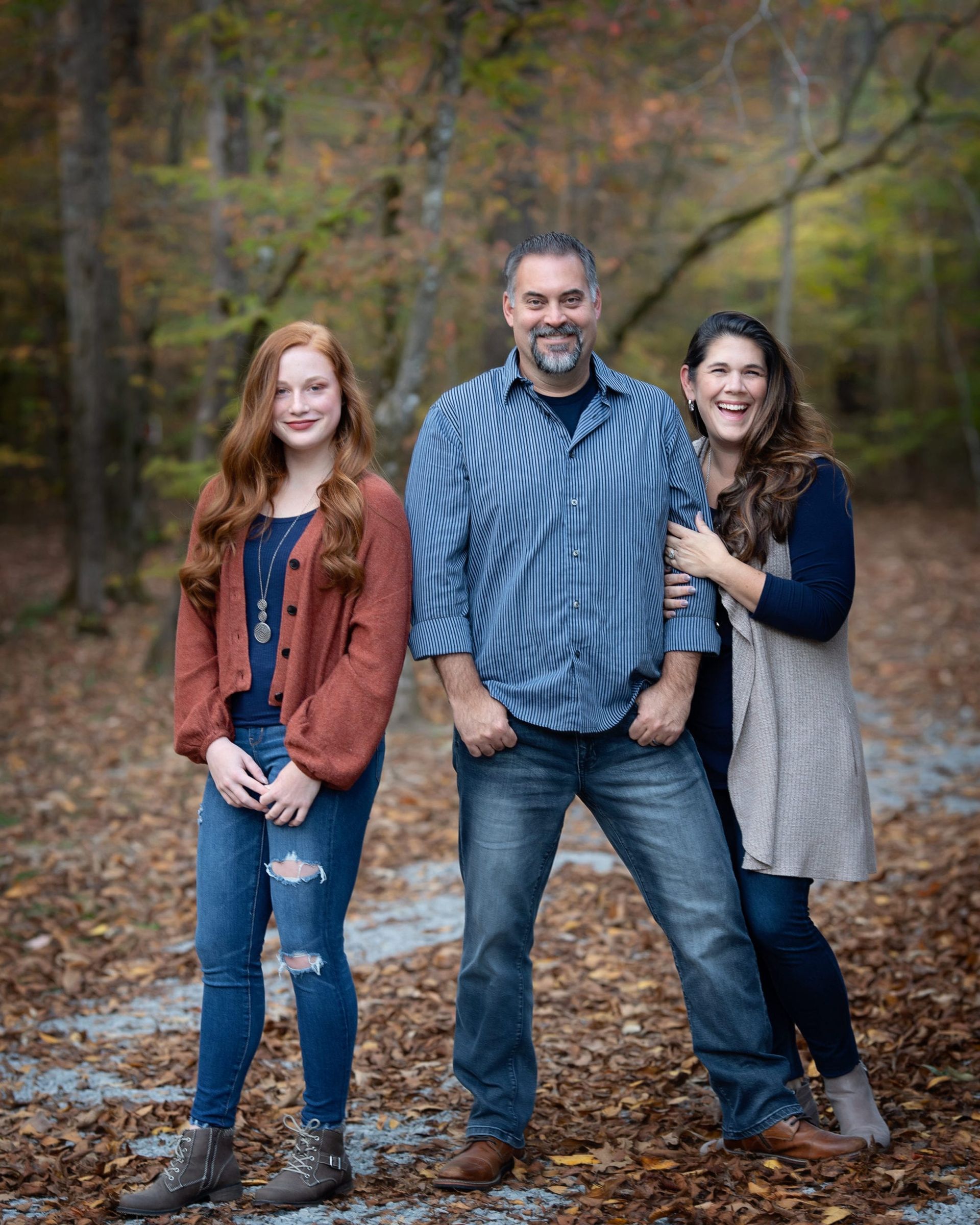 Family of three posing outdoors: red-haired daughter, smiling parents, fall foliage background.