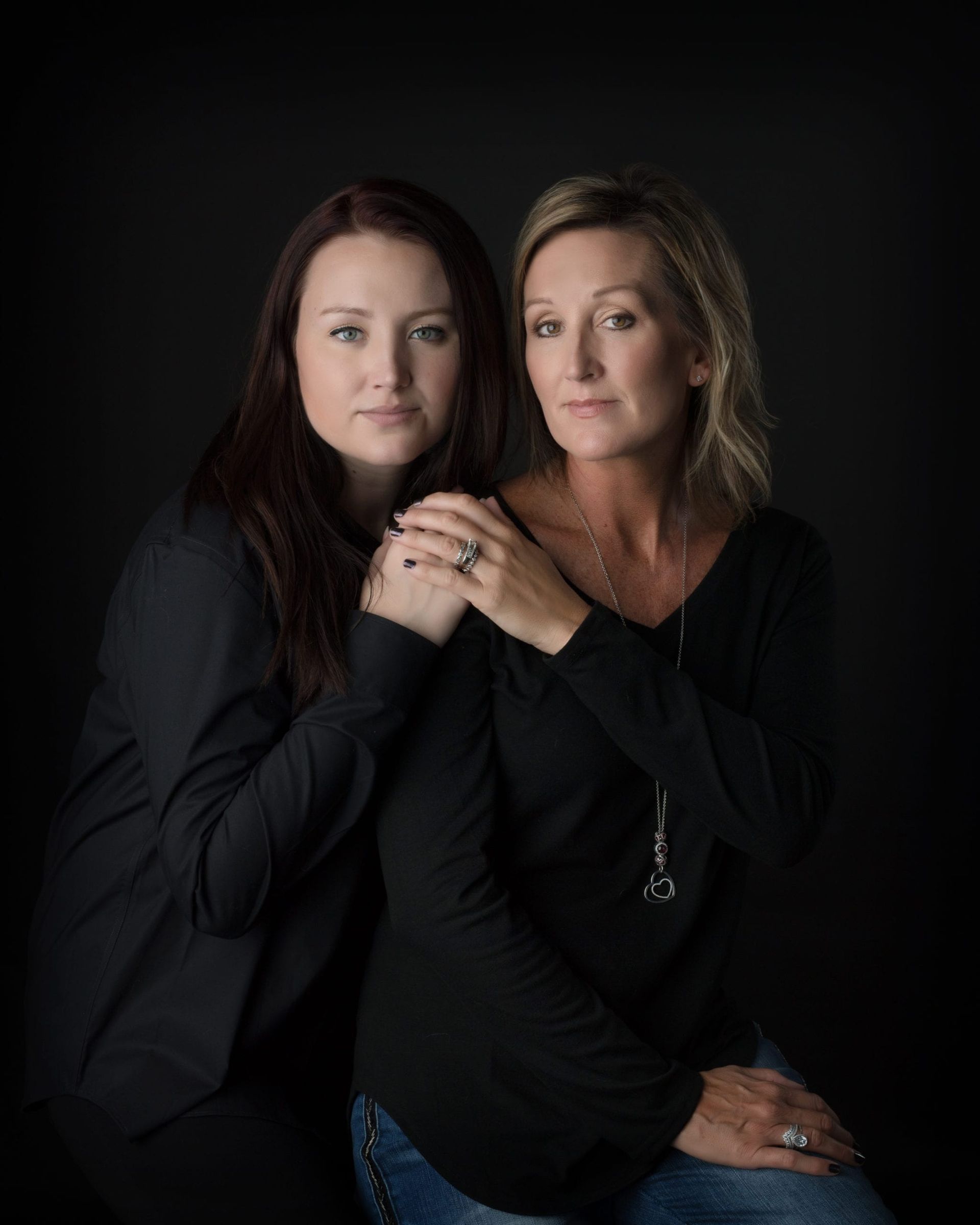 Two women posing together against a black background; one has her hand on the other's shoulder.