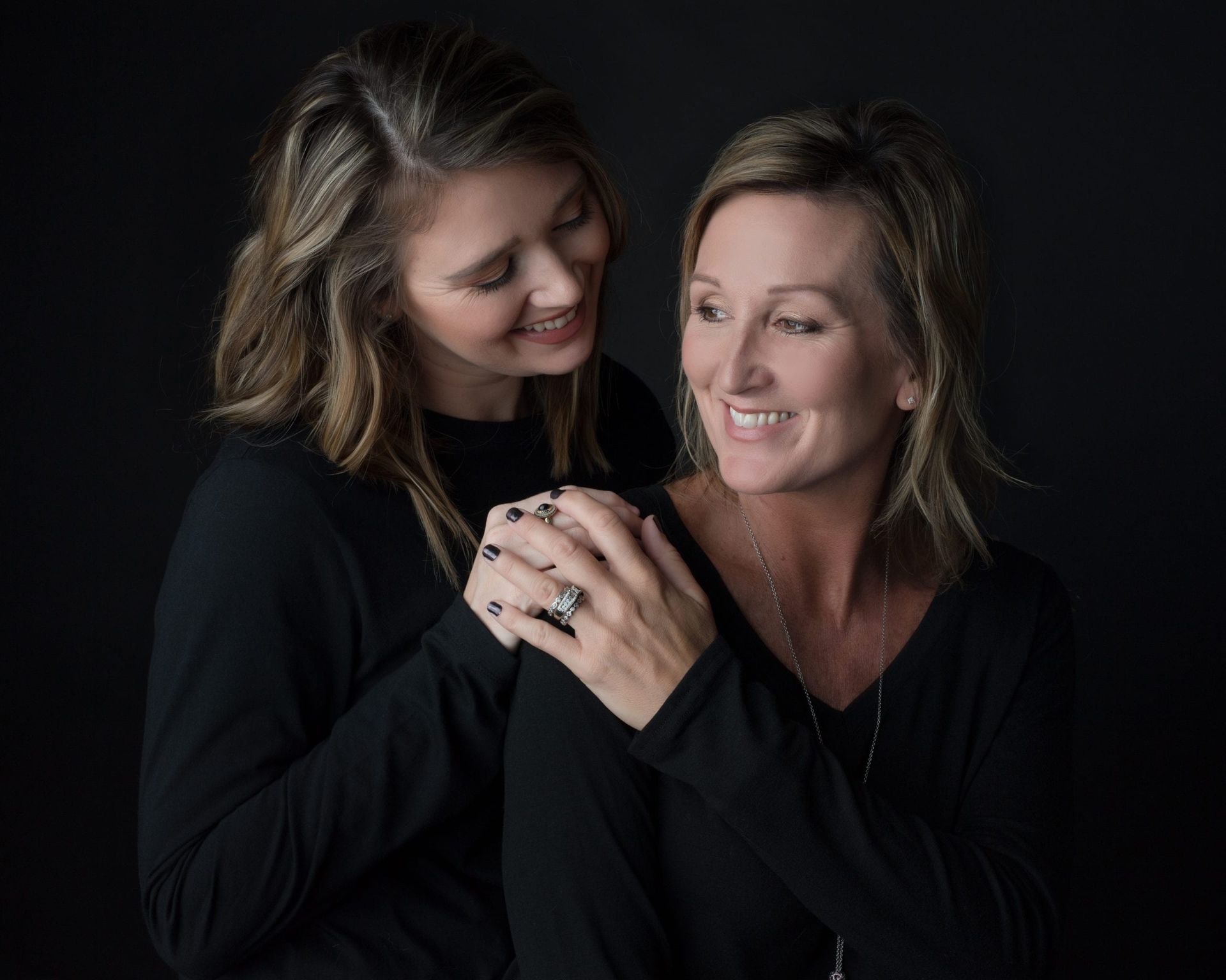 Mother and daughter smiling, posing close together, wearing black tops, against a dark background.