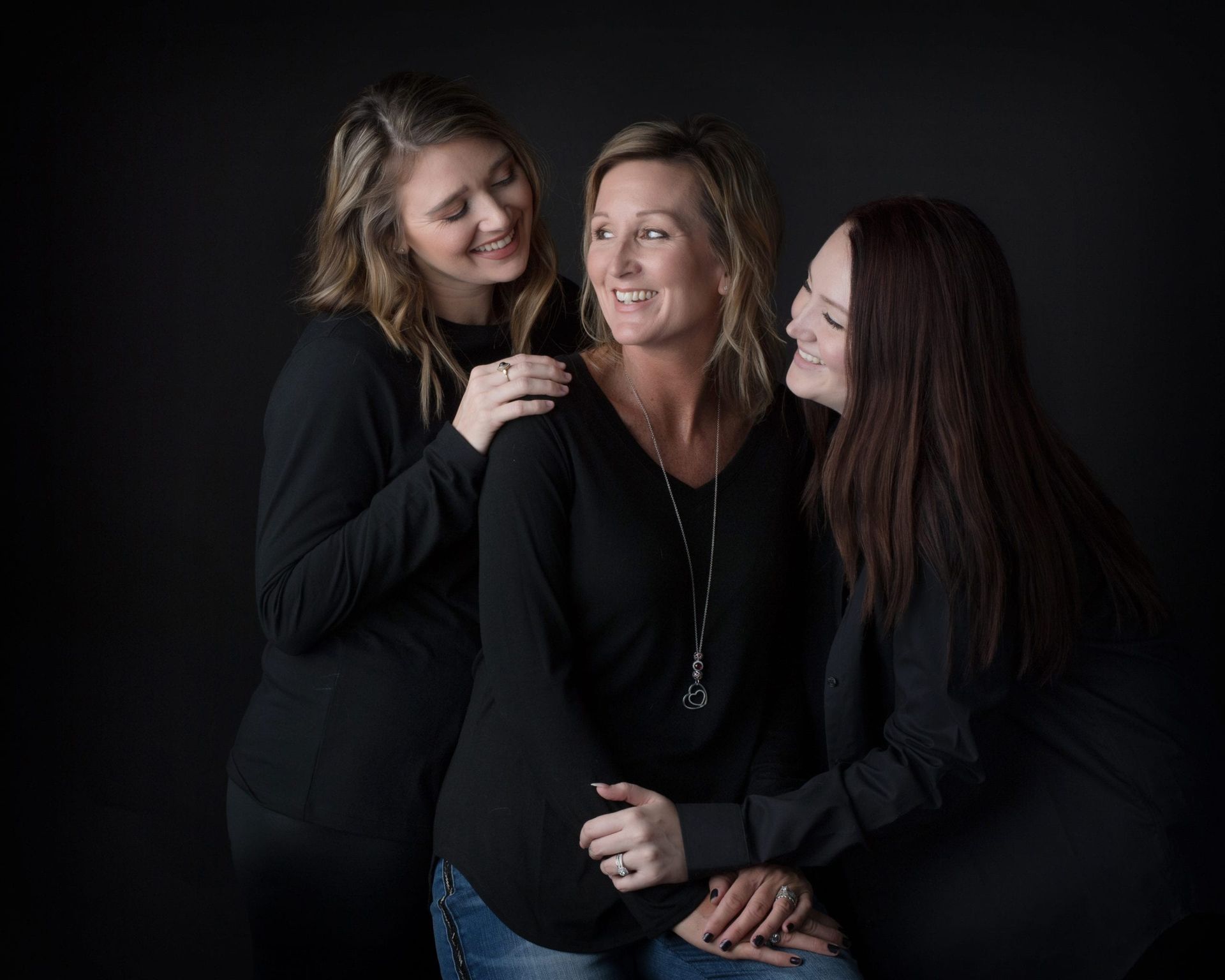 Mother and two daughters in black shirts, smiling, embracing against a dark backdrop.