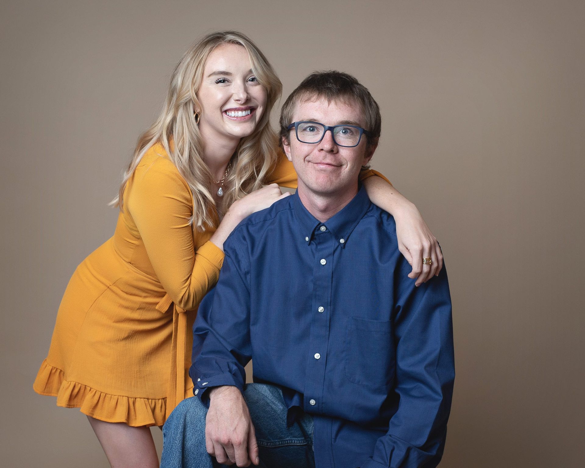 Young woman in yellow dress hugs man in blue shirt, both smiling, against neutral background.