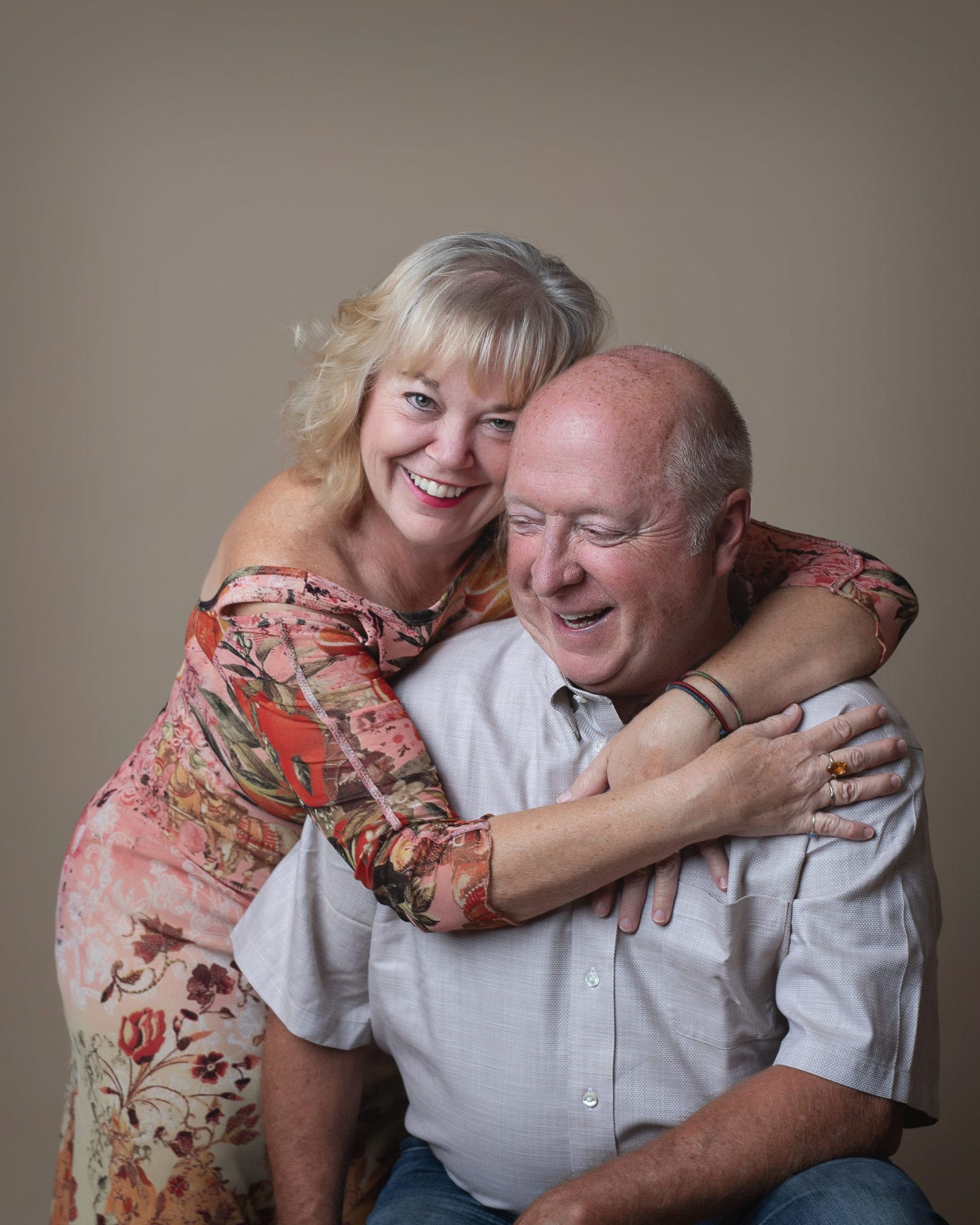 Smiling woman hugs bald man, both appear joyful, against a neutral backdrop.