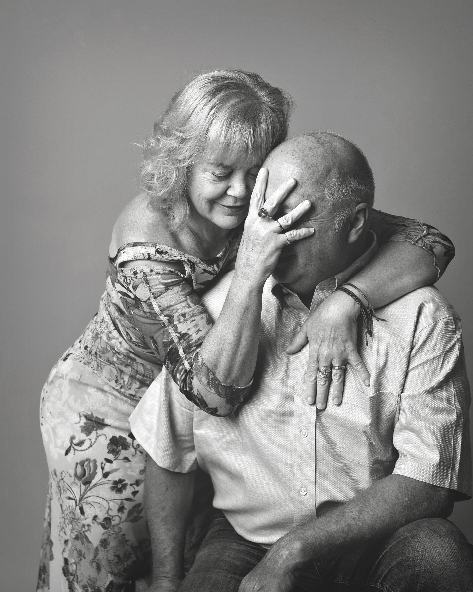 Woman comforts man; she hugs him, covering his eyes. Black and white.
