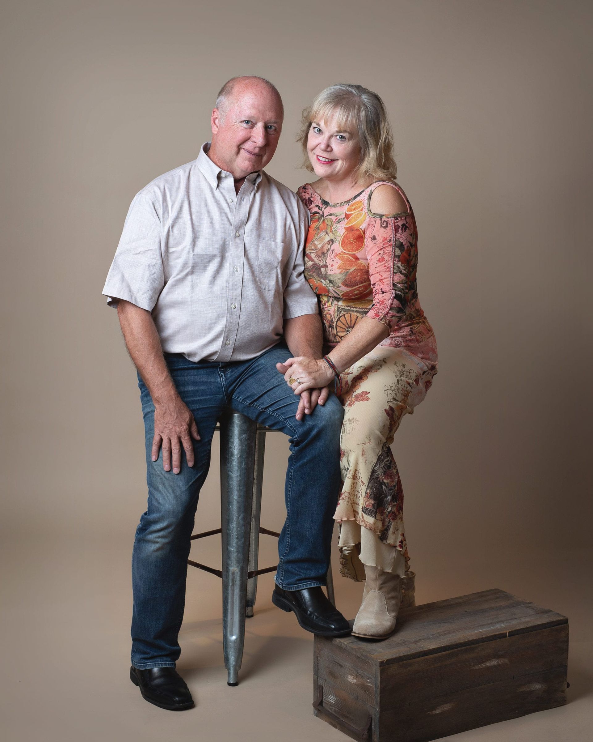 Man sits on a stool, smiling. Woman stands beside him on a box, smiling. Beige background.