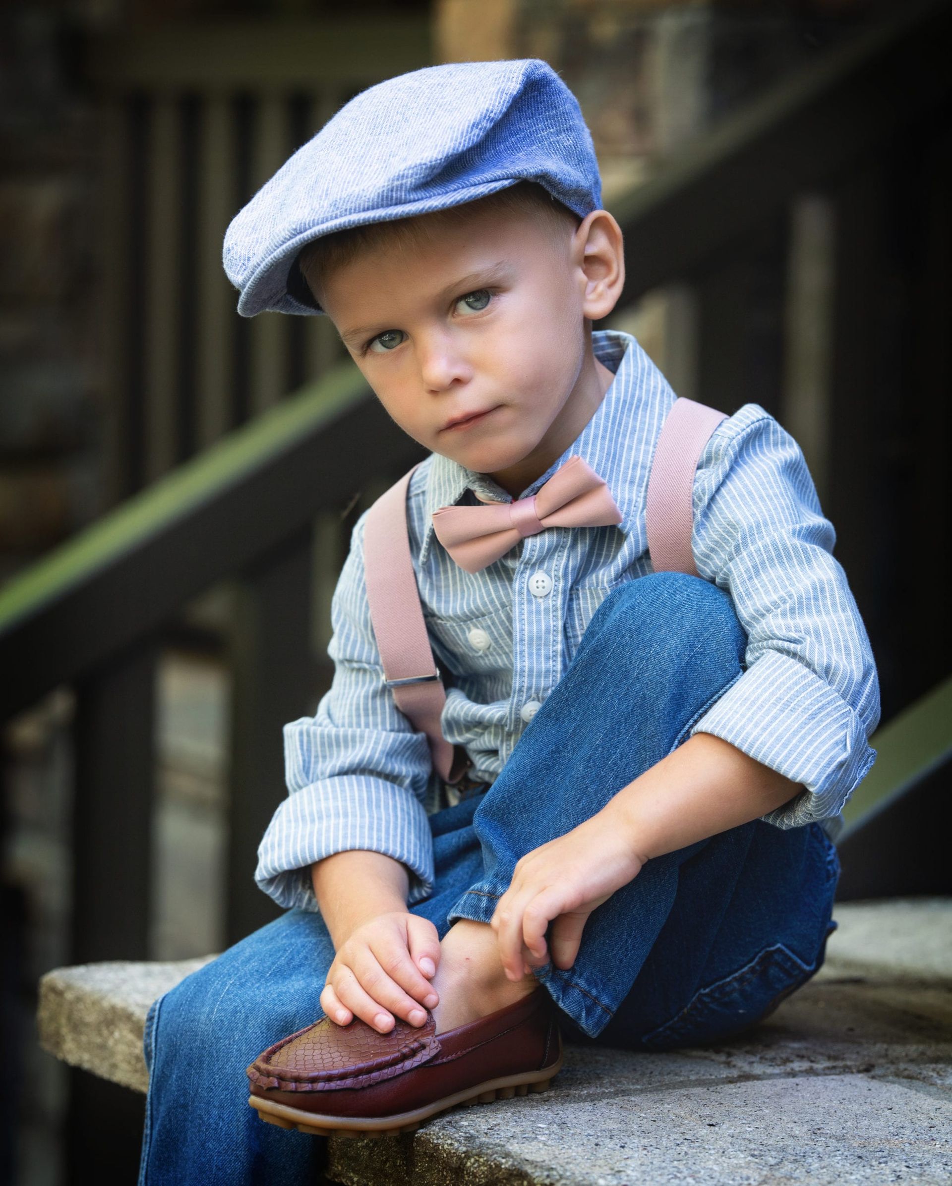 Boy in a blue cap, striped shirt, jeans, suspenders, and bow tie, sitting and looking down.