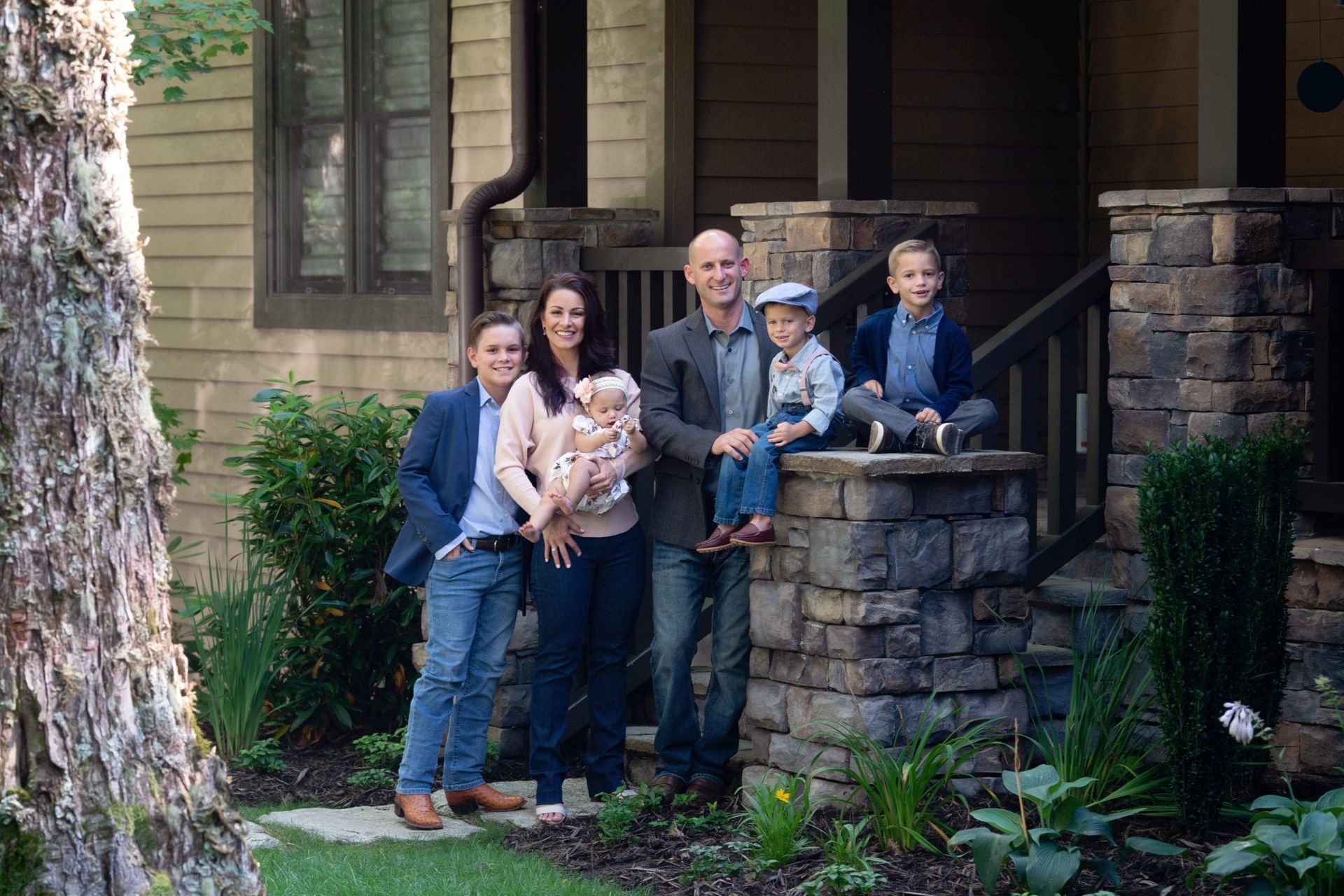 Family of six poses in front of their home; two boys sit, one held. Beige exterior, stone columns.