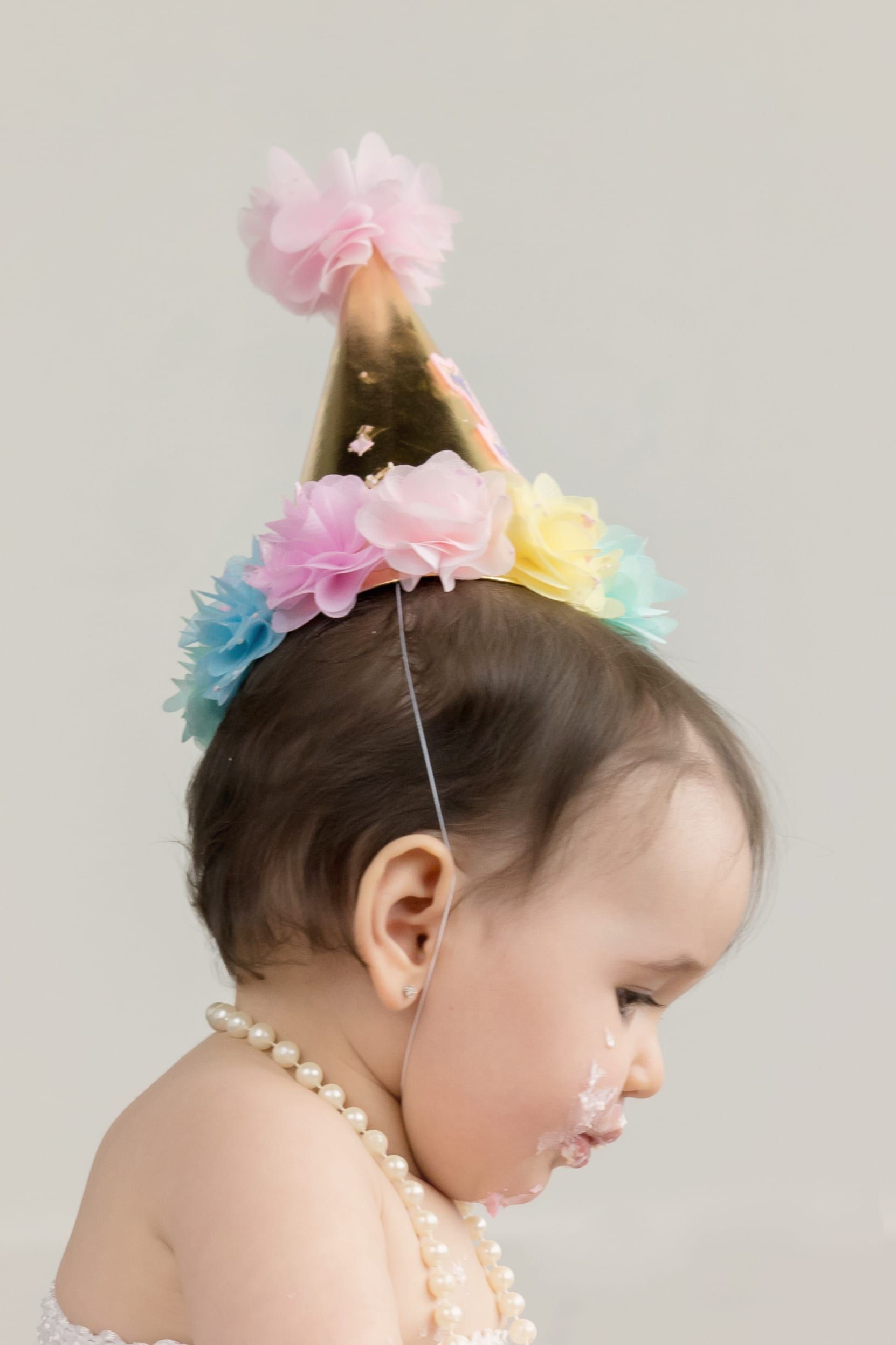 Baby with a birthday party hat and pearl necklace, covered in cake.