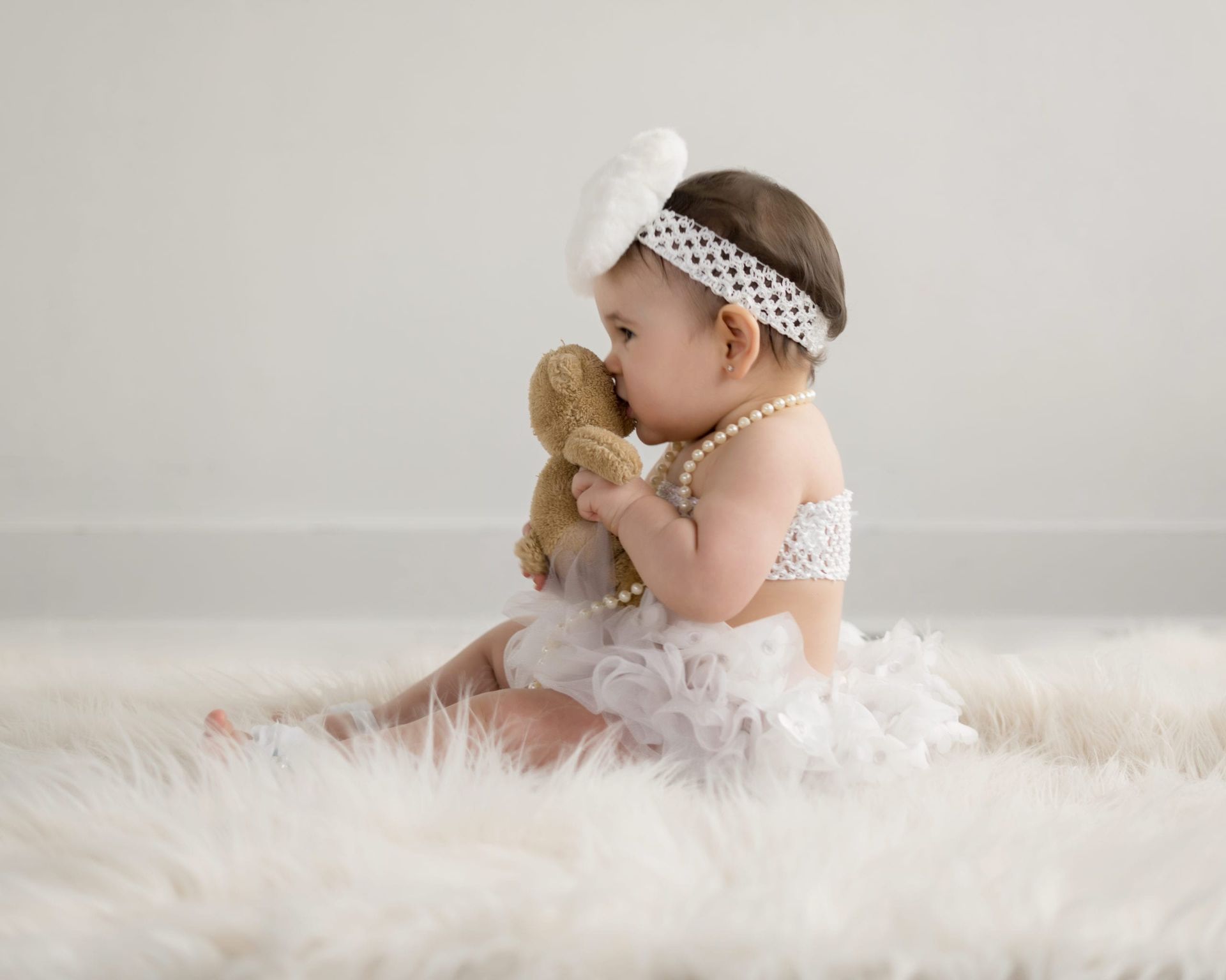 Baby in white lace outfit, headband, and pearls, kissing a teddy bear on a fluffy white rug.