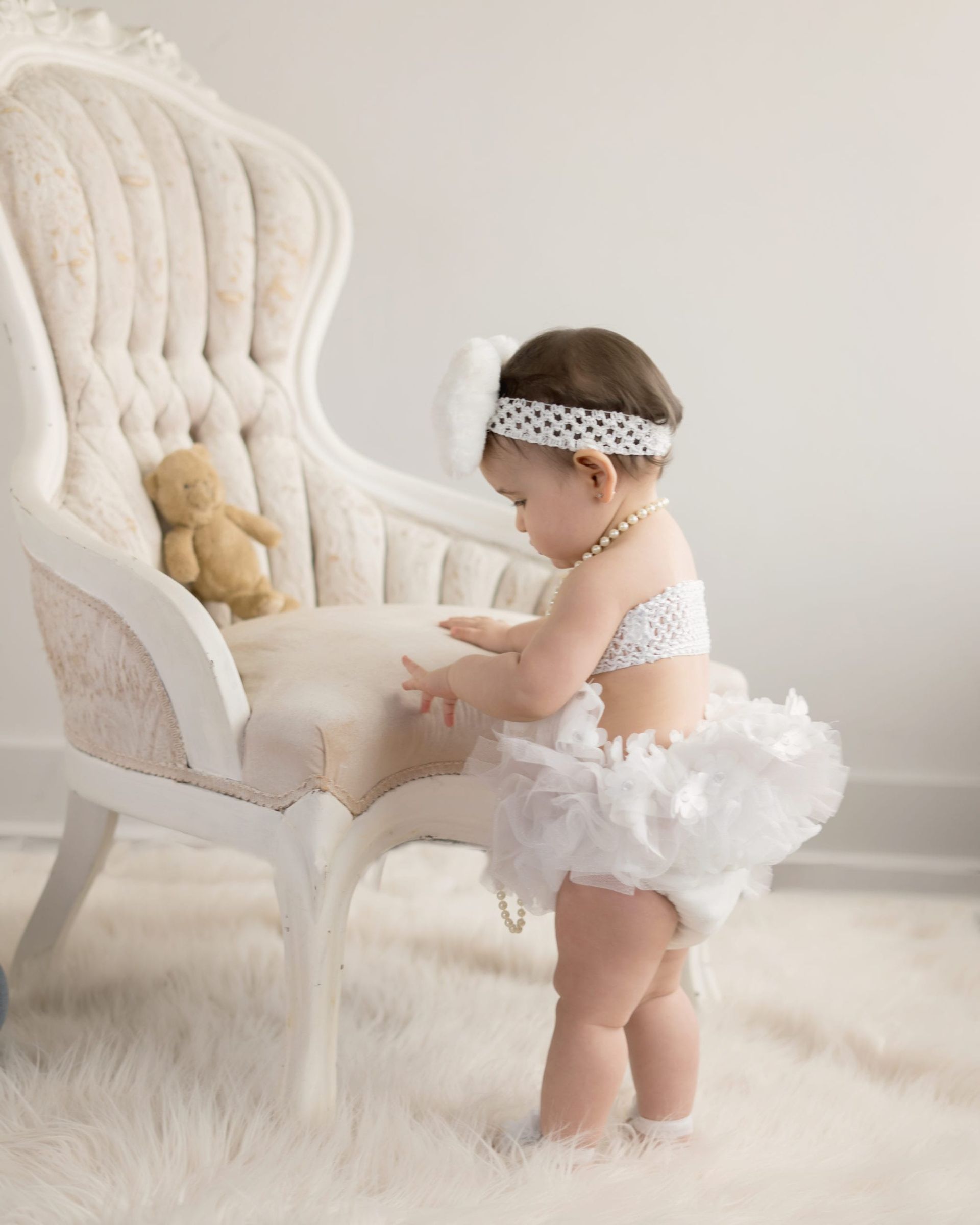 Baby girl in a tutu and headband, standing near a cream-colored chair and a teddy bear on a white rug.