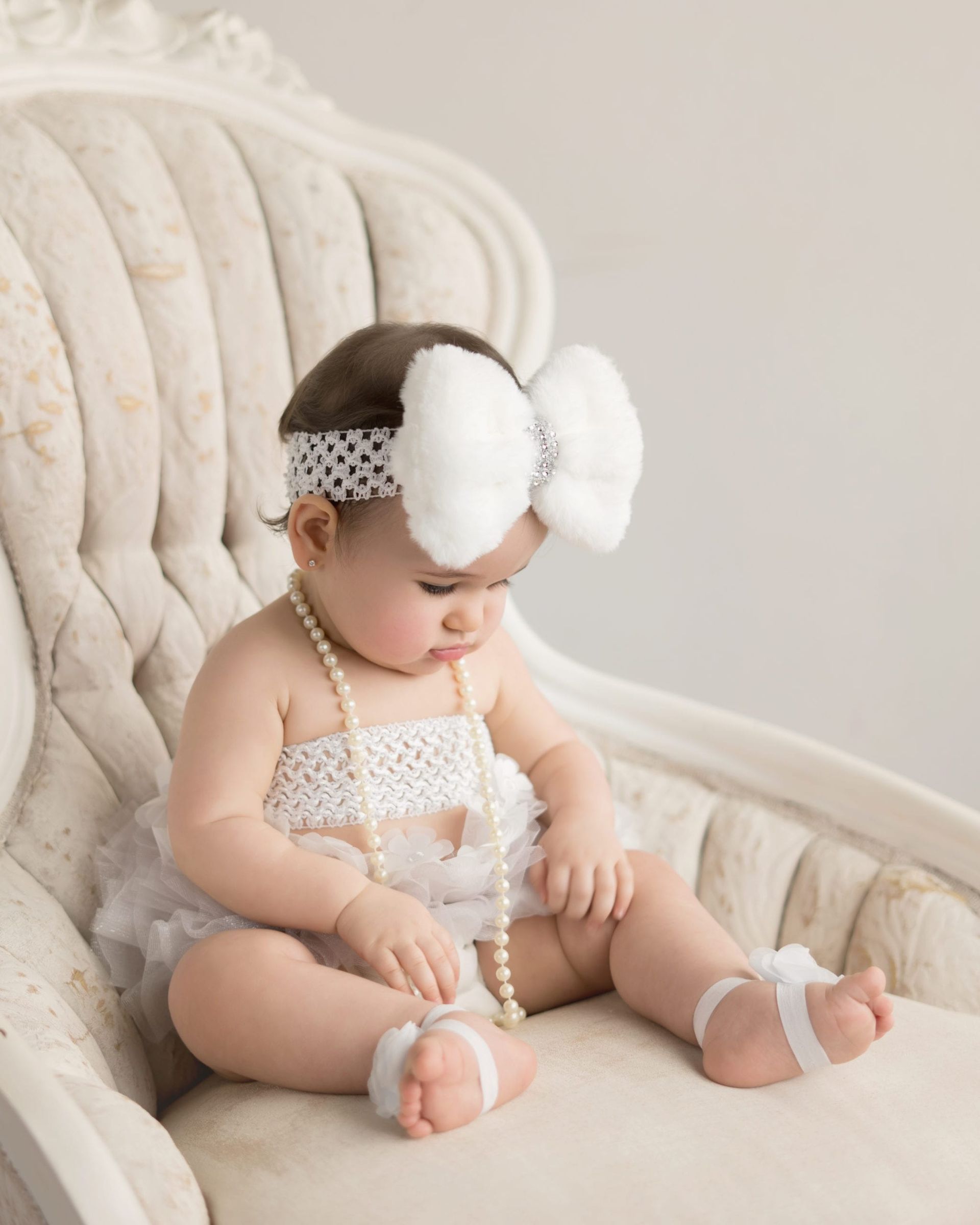 Baby girl in white outfit and bow, sitting on a vintage chair.