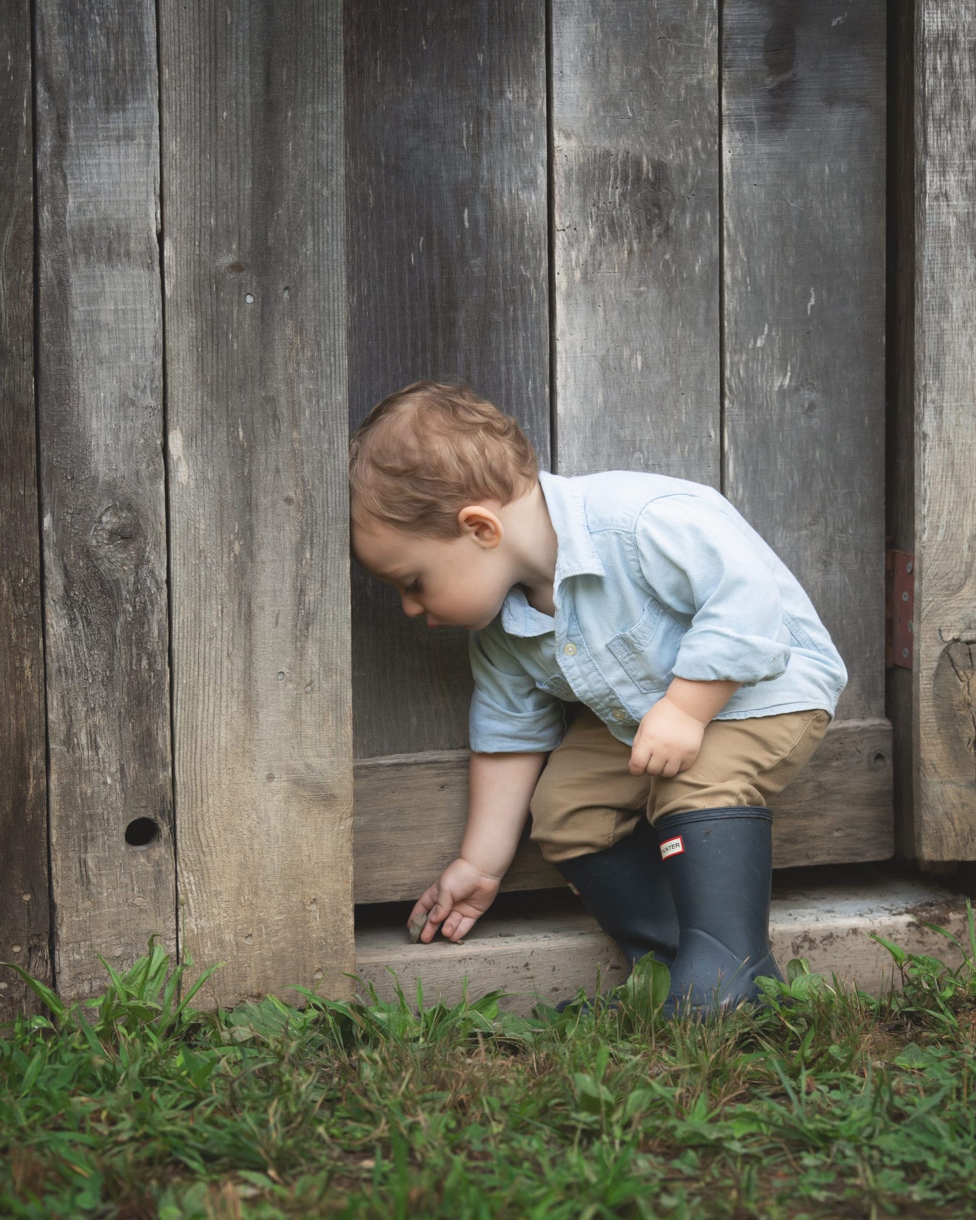 Young child in blue shirt and boots examines something on a concrete step next to weathered wooden siding.
