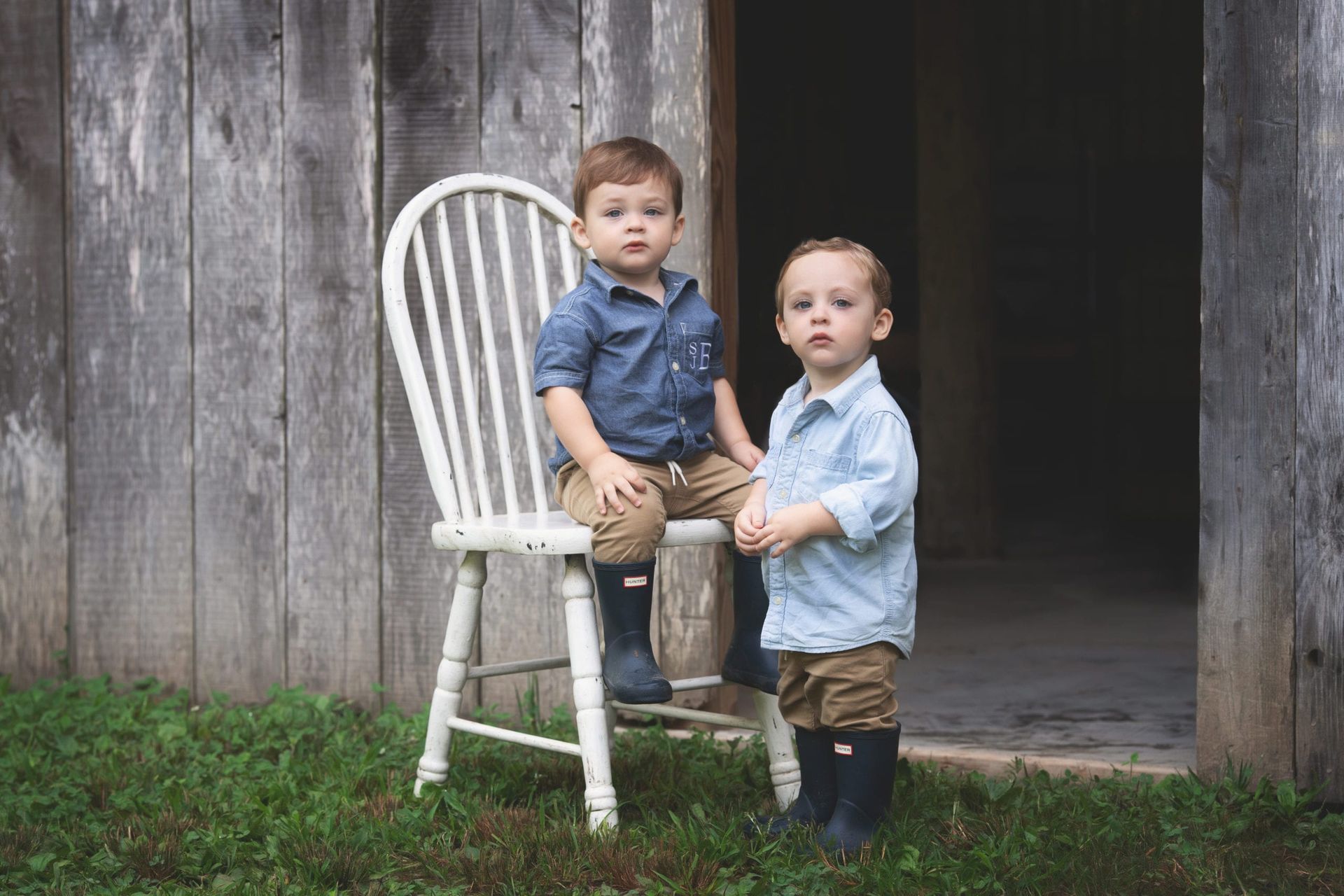 Two young boys pose next to a barn. One sits in a white chair, the other stands. Both wear blue shirts, brown pants, and boots.