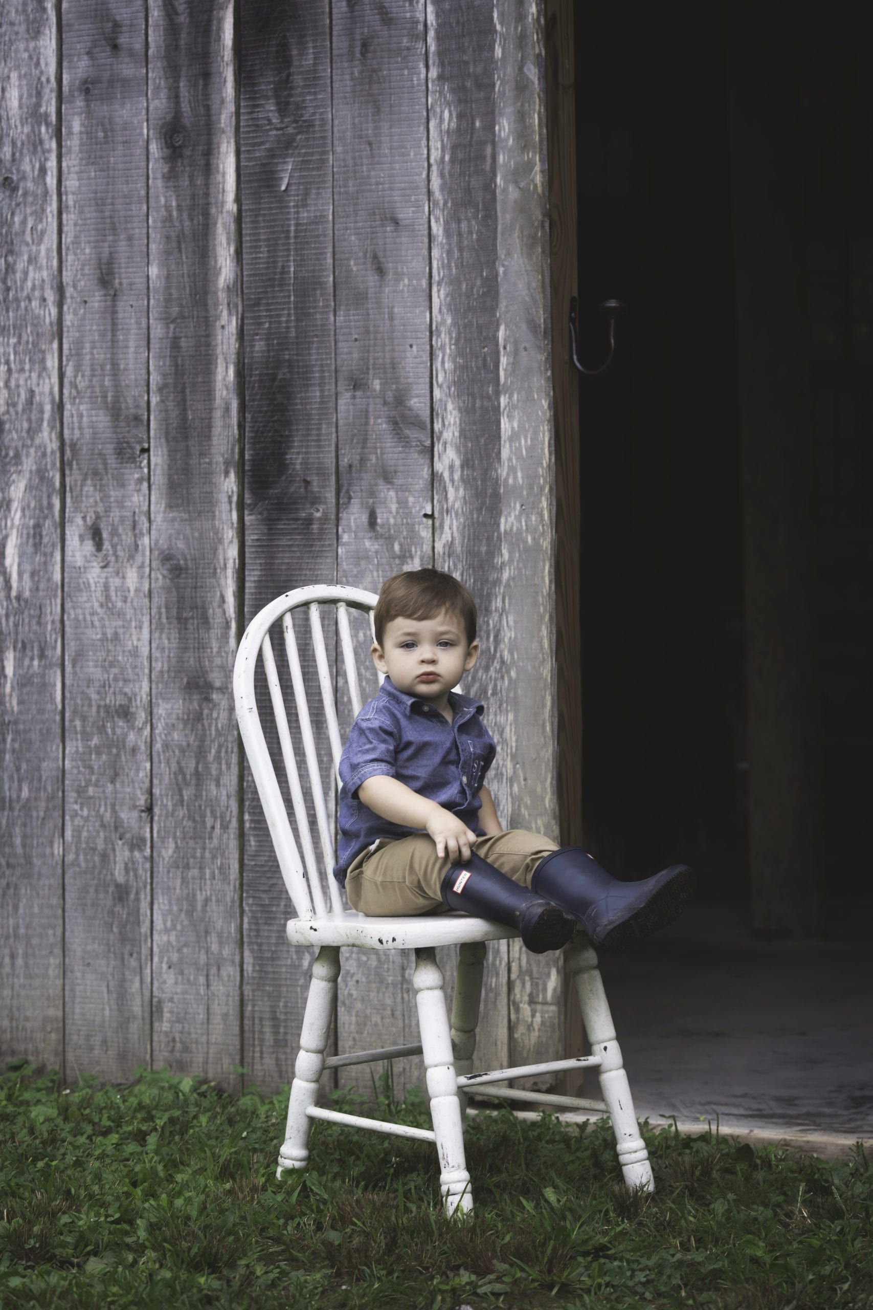 Boy in blue shirt and boots sits on a white chair in front of a wooden barn.