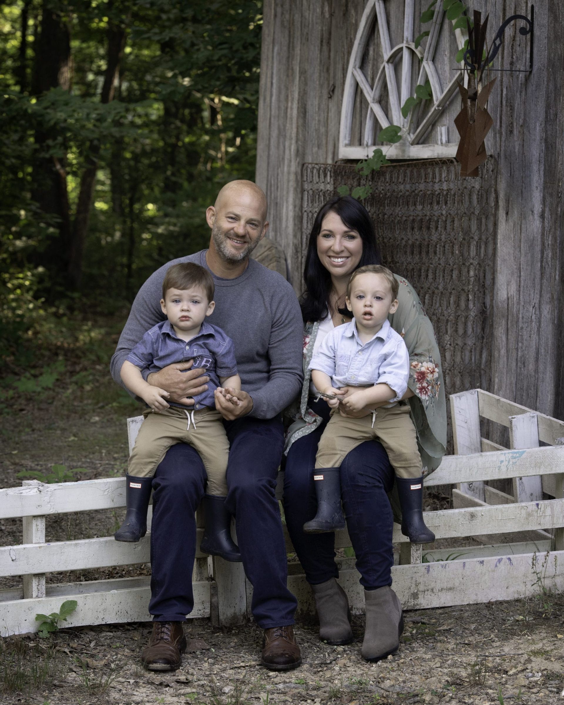 Family of four sits on a fence; the parents hold their two children. Outdoors, near a wooden structure.