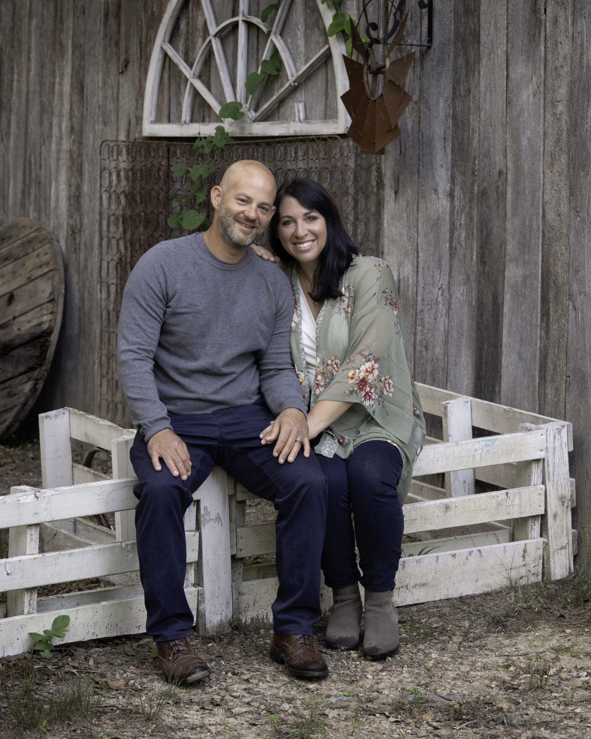 Couple smiling, sitting together on a white fence in front of a rustic wooden wall.