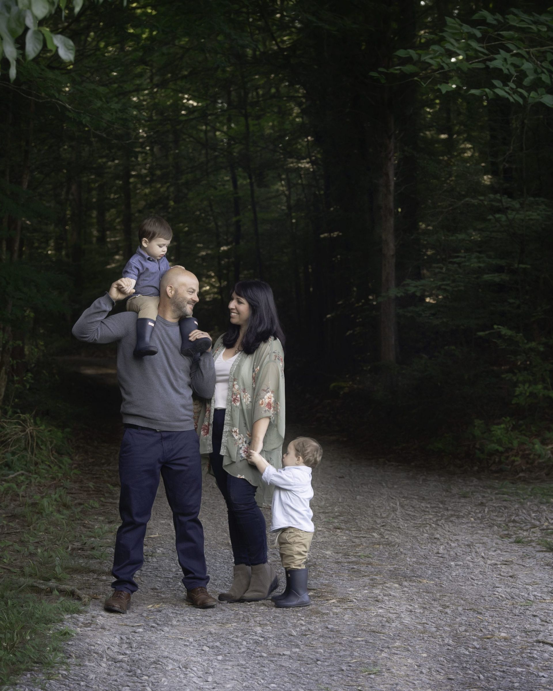 Family of four on a gravel path in a forest; father with son on shoulders, mother holding son's hand.