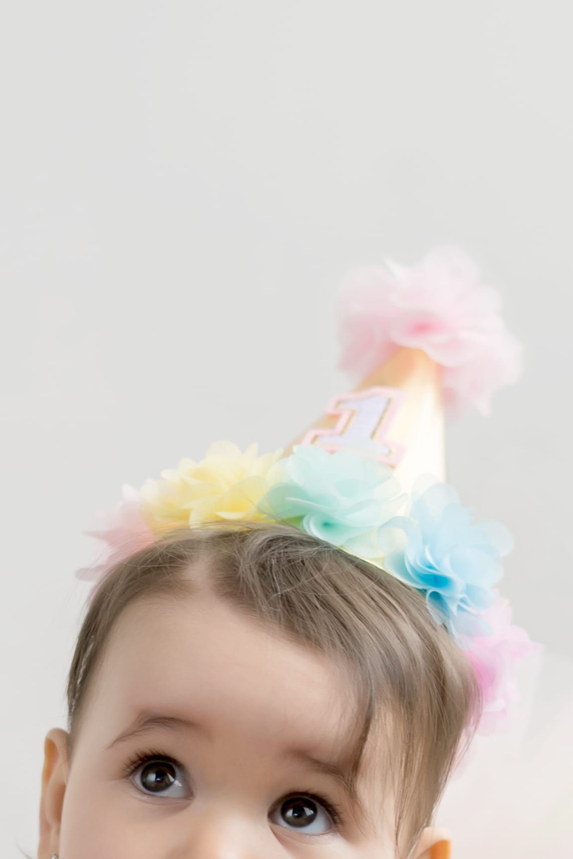 Baby wearing a pastel-colored birthday party hat, looking upward with large eyes.