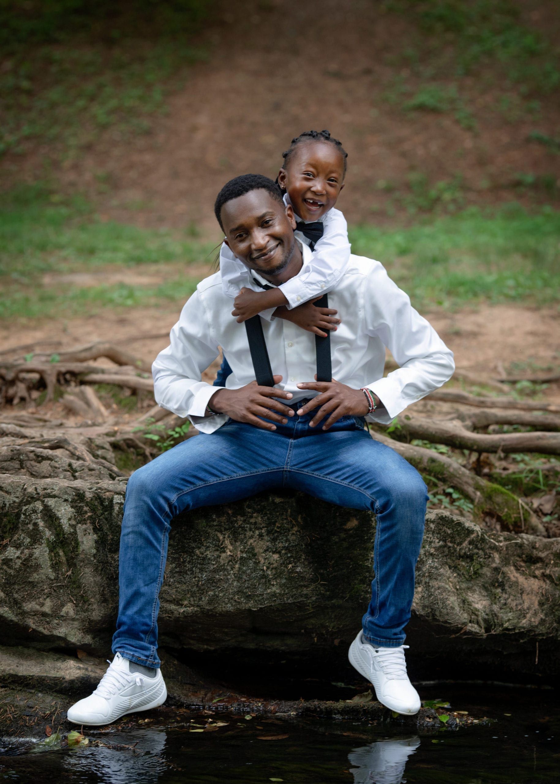 Man and child smiling, embracing, outside. Man sits on rock; child is on his back. Green foliage in background.