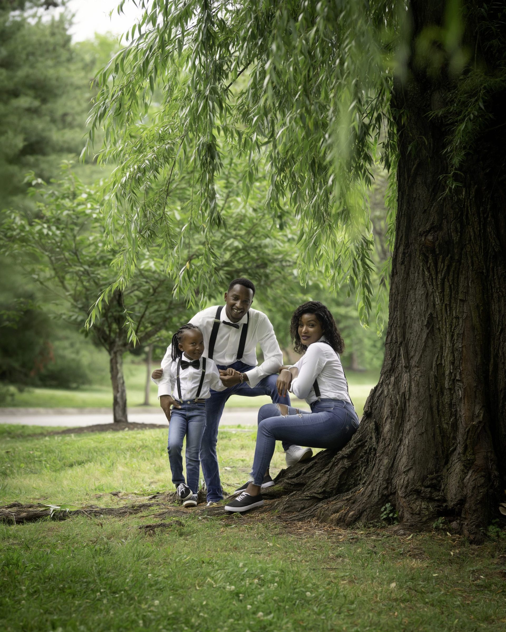 Family of three pose under a tree in a park. They wear white tops, suspenders, and jeans, smiling.
