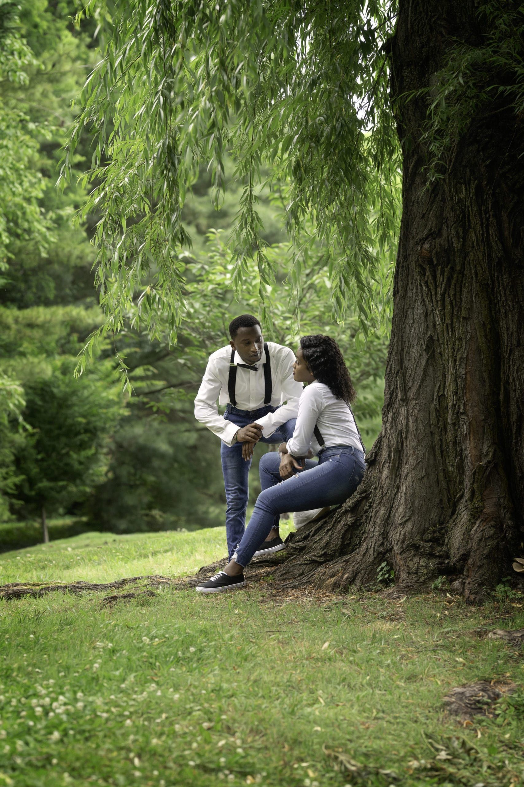 Man and woman by a tree in a park. Man holds woman's hand. Both are smiling.