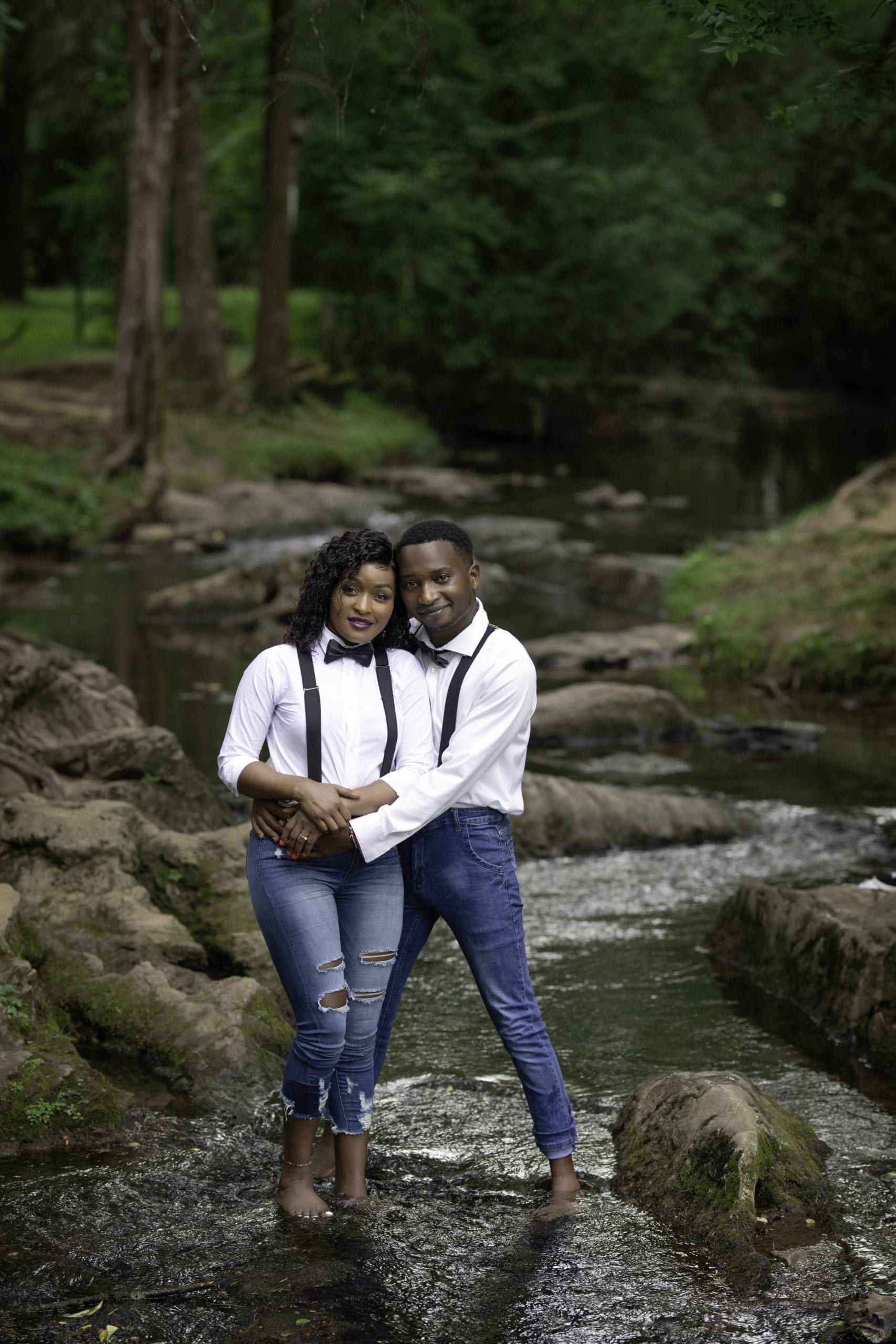 Couple in white shirts, bow ties, and jeans stands in a stream, smiling, embracing in a wooded area.
