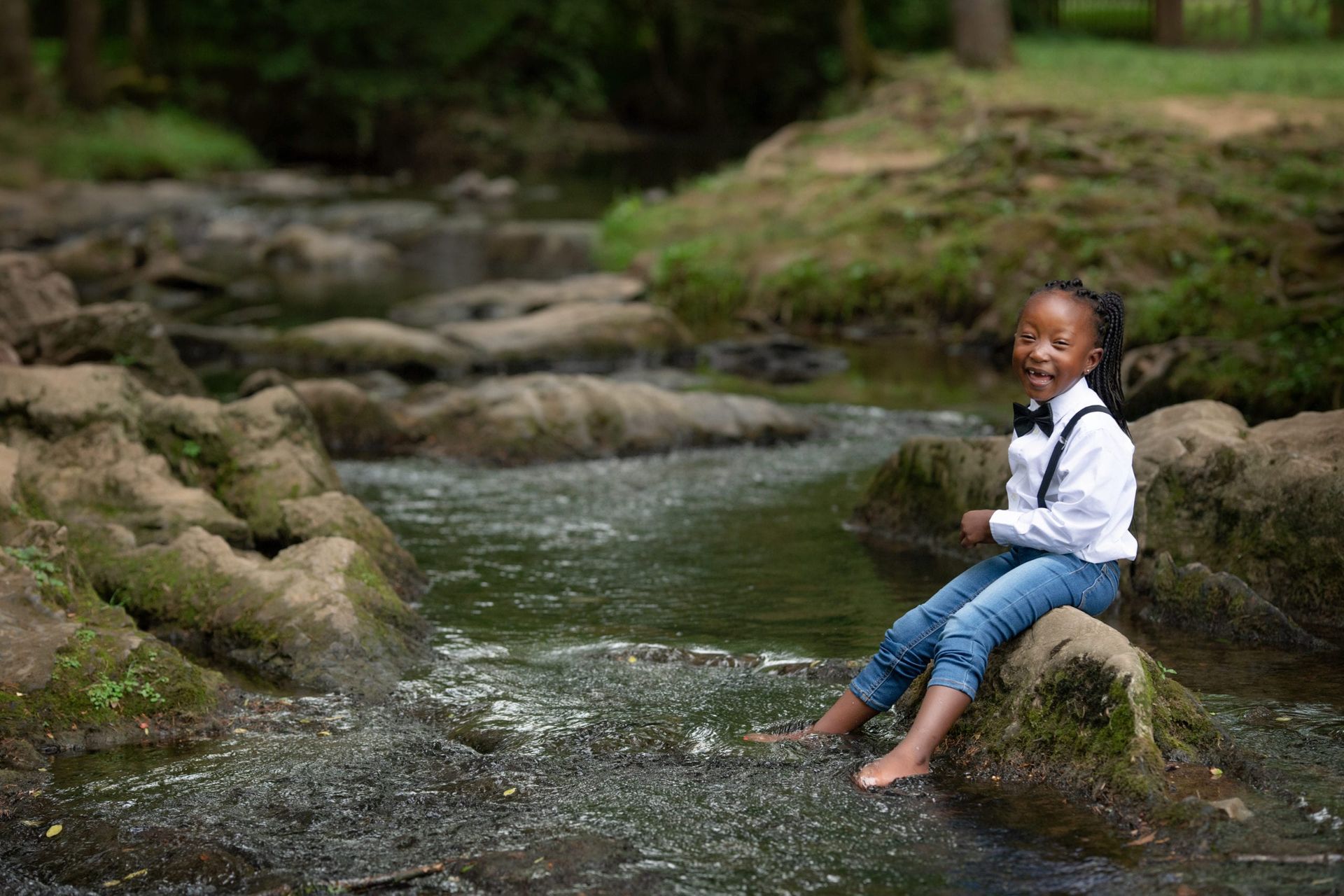 Young Black child with a bow tie sits on a rock in a stream, smiling, with bare feet in the water.