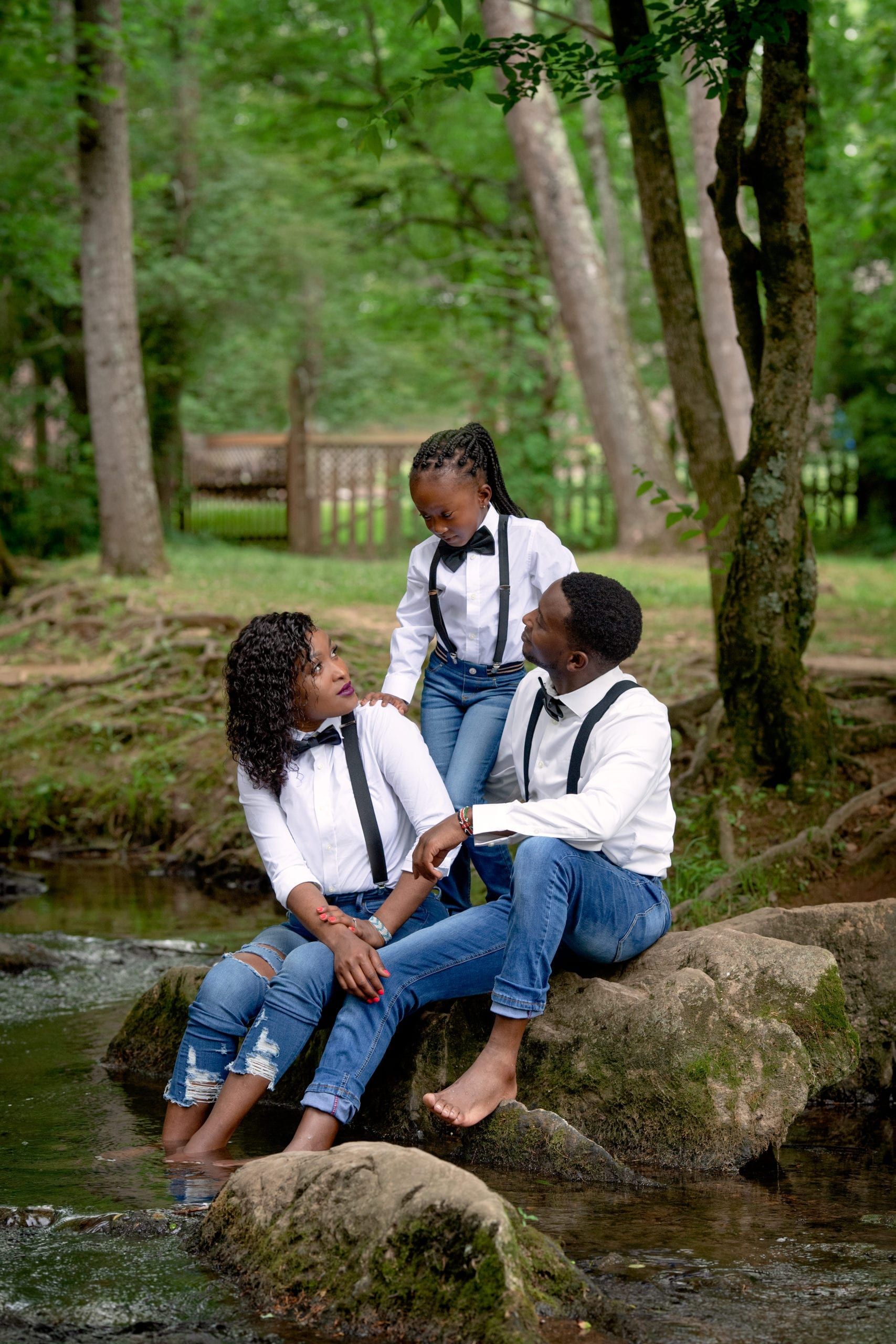 Family of three smiling, sitting on rocks in a creek, wearing white shirts, blue jeans, and bow ties, in a wooded area.