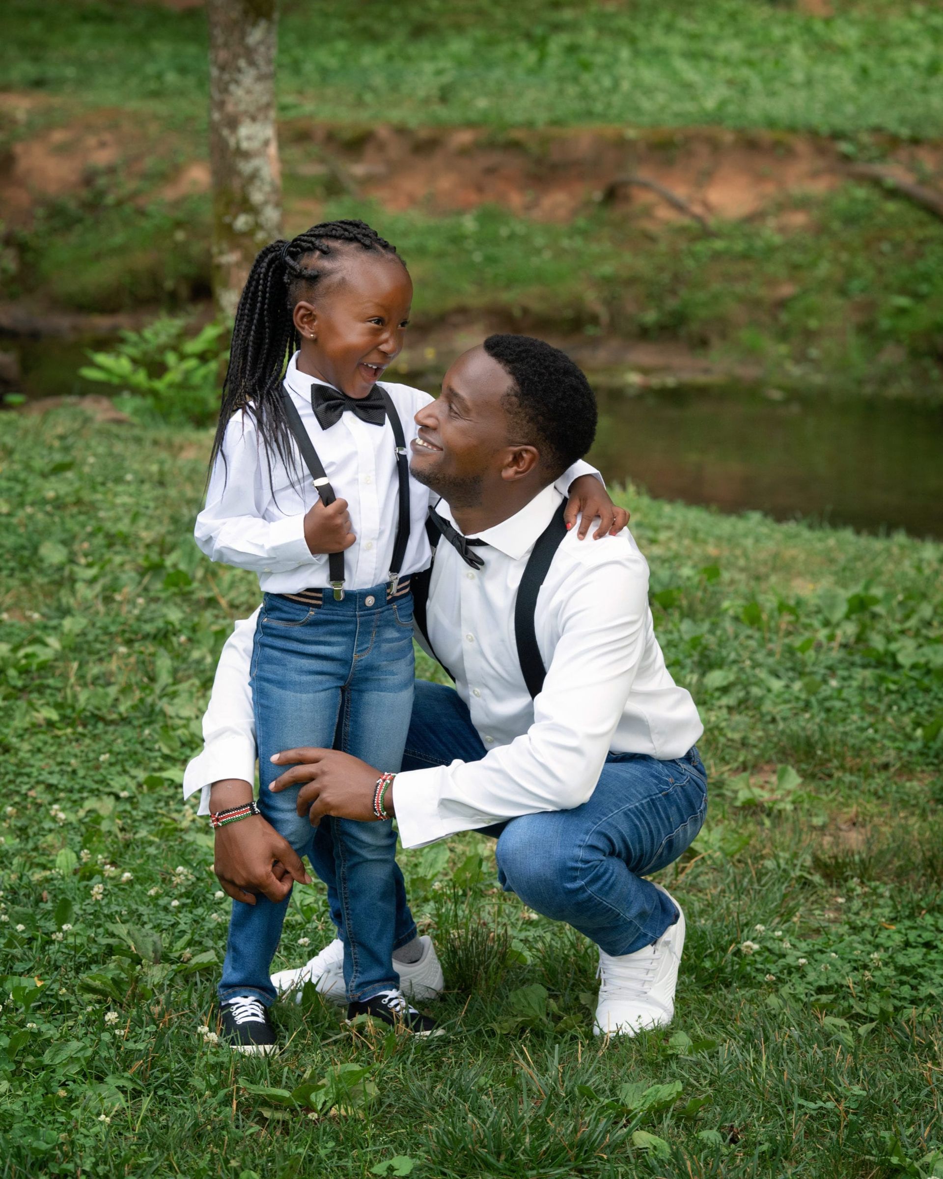 Father and daughter smiling at each other. They wear bow ties and jeans, in a park setting.