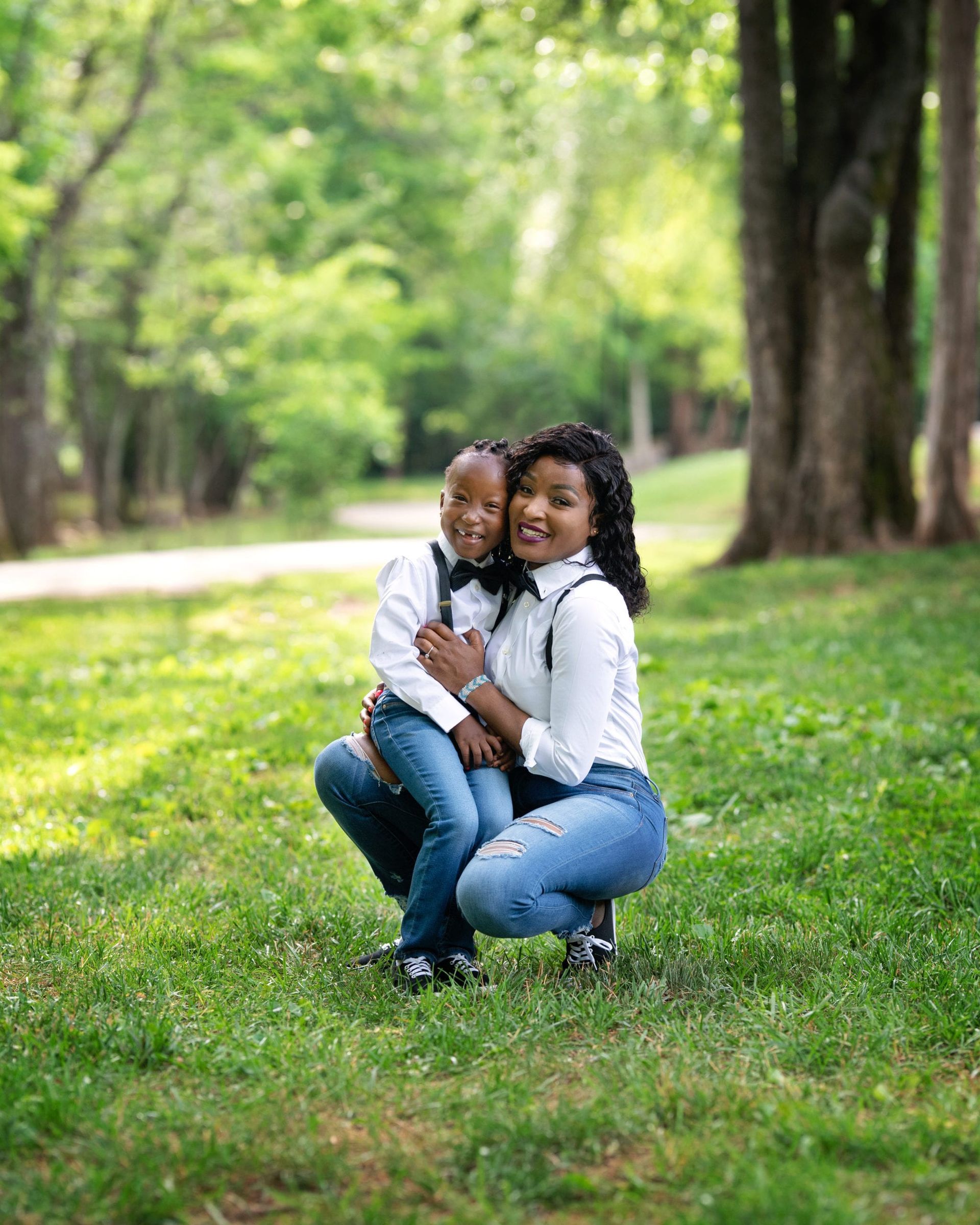 Mother and child pose in a park. They wear white shirts and jeans, smiling. Sunlight streams through the trees.