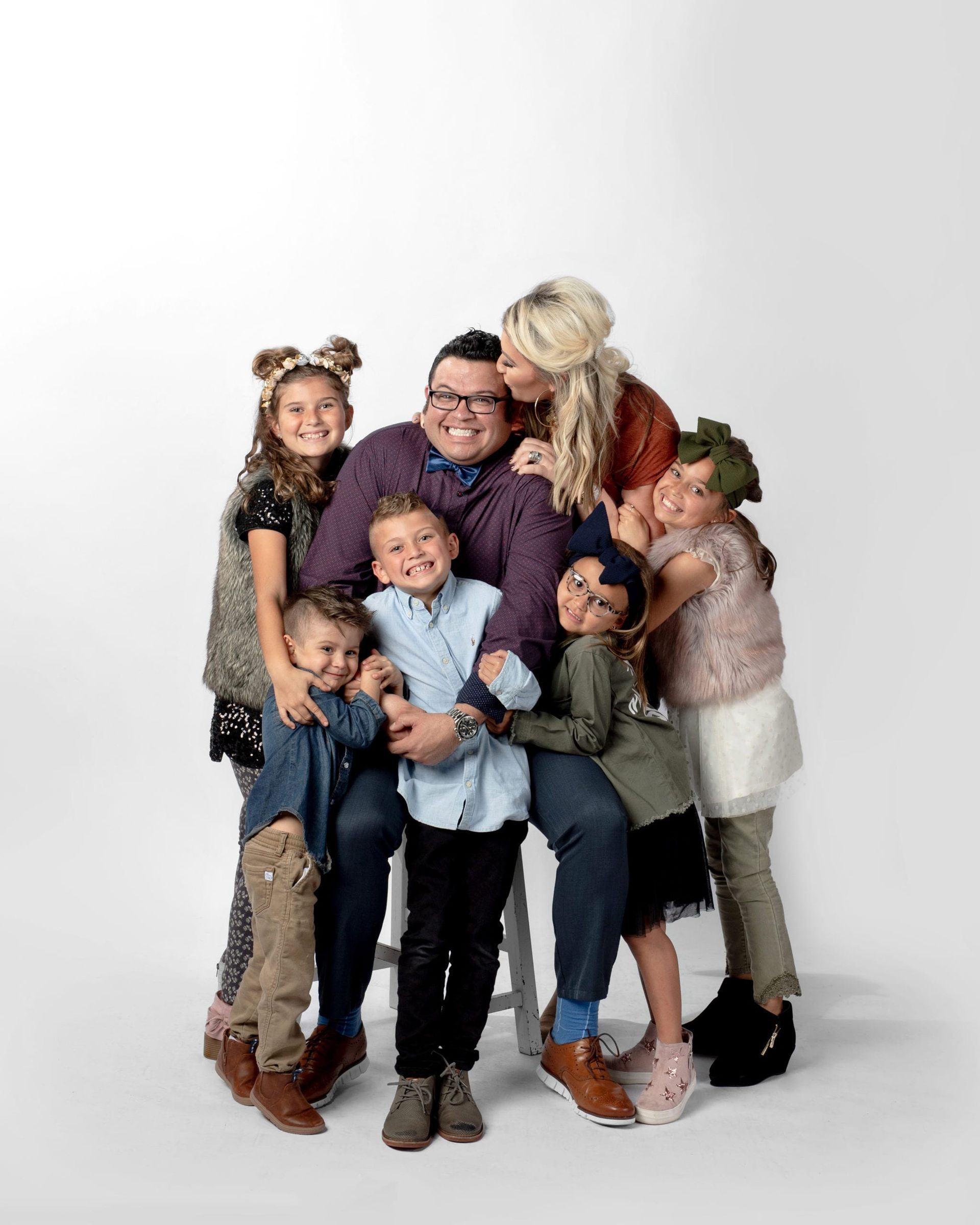 Man surrounded by smiling children and woman kissing him, all in a studio setting.