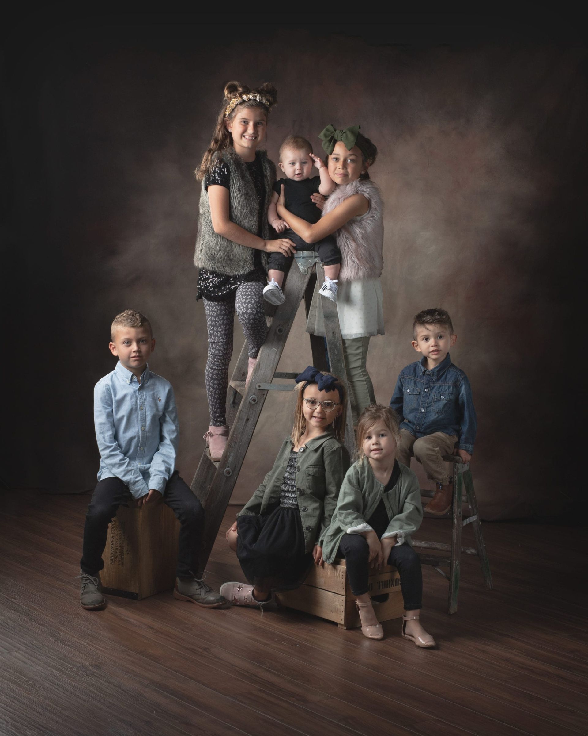 Group of seven children posing on and around a ladder, dressed in whimsical outfits against a dark backdrop.