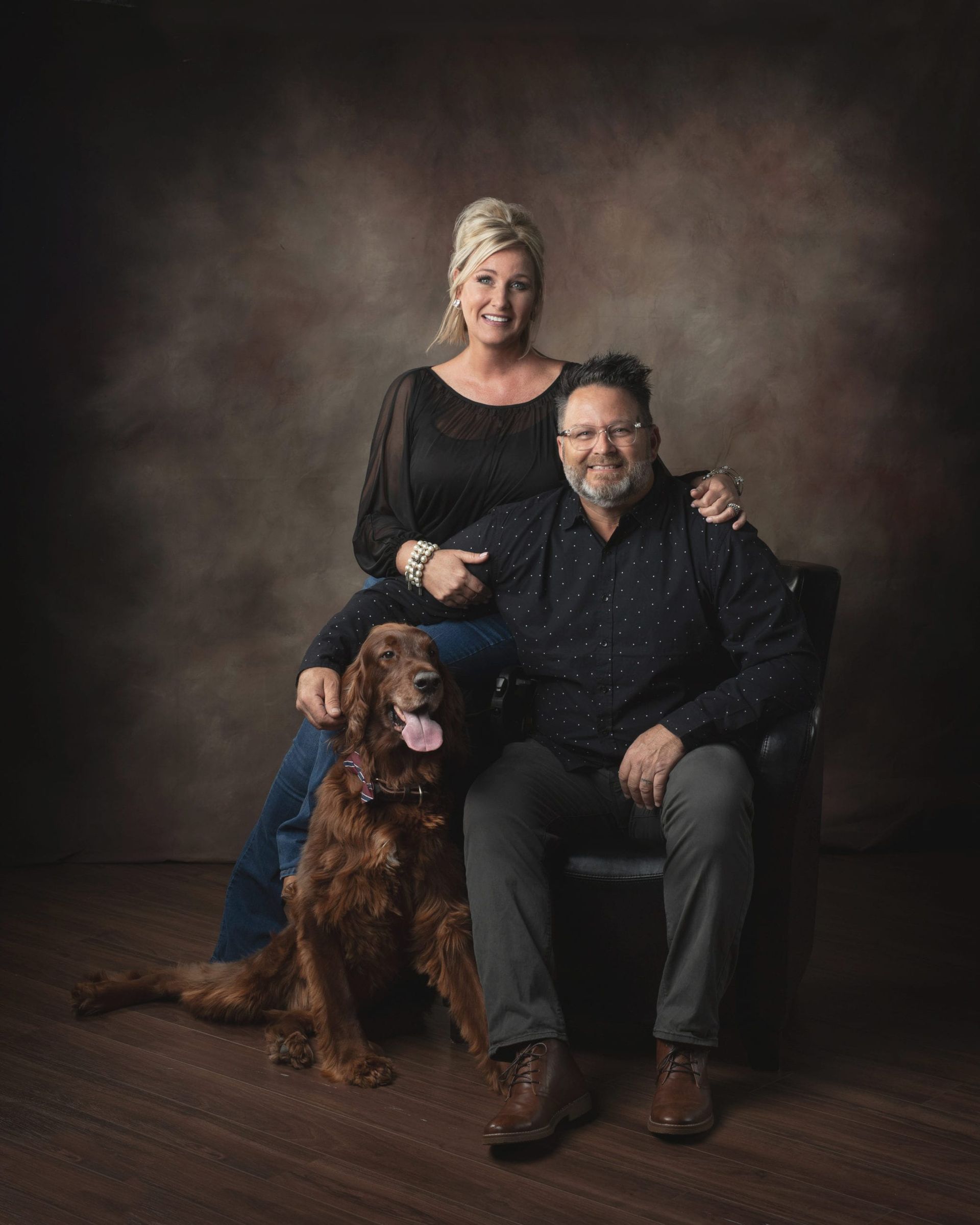 Couple and dog pose in studio, man seated, woman behind him, brown dog sits by.